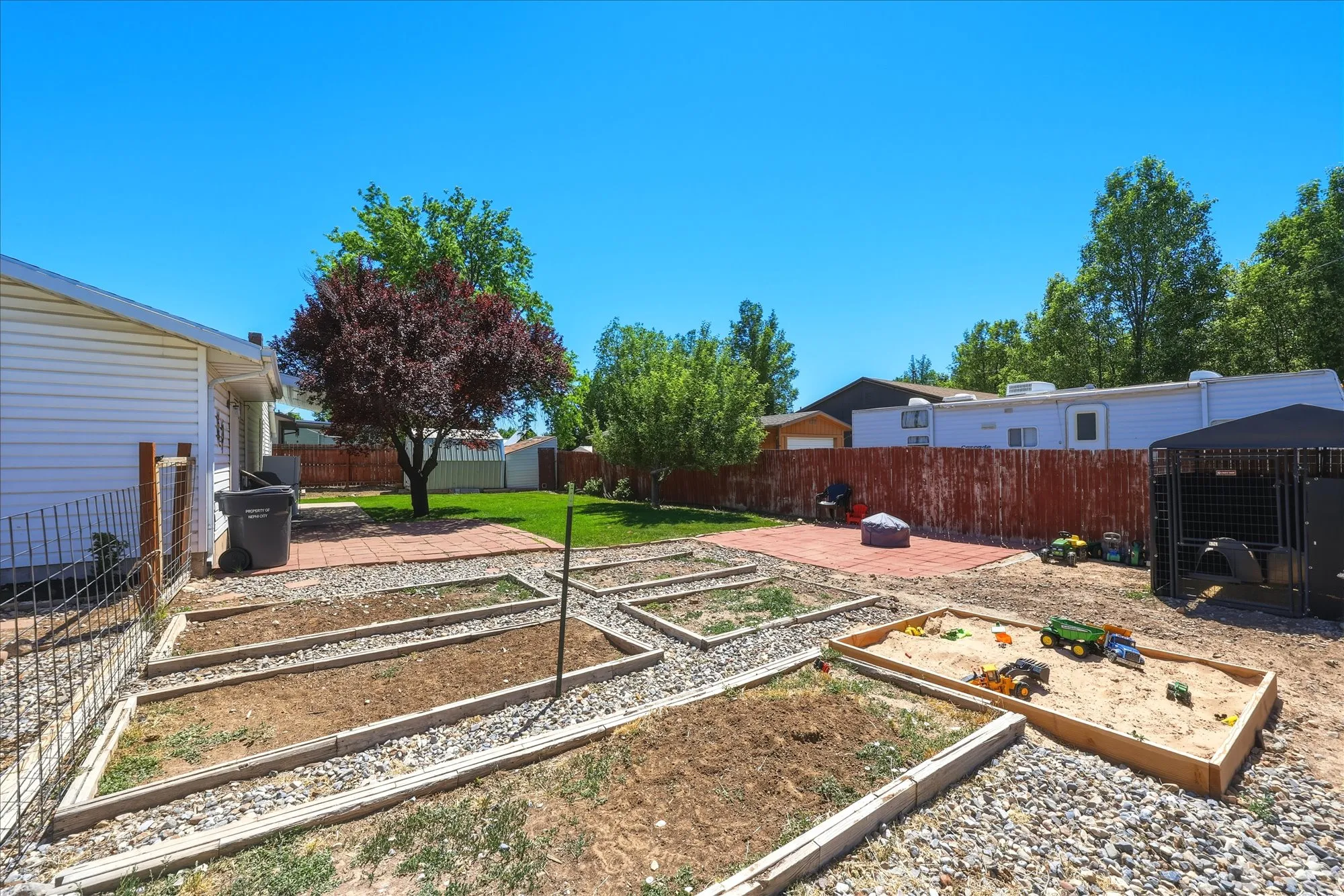 Expansive backyard featuring multiple raised garden beds, a dedicated sandbox area, and a large paved patio