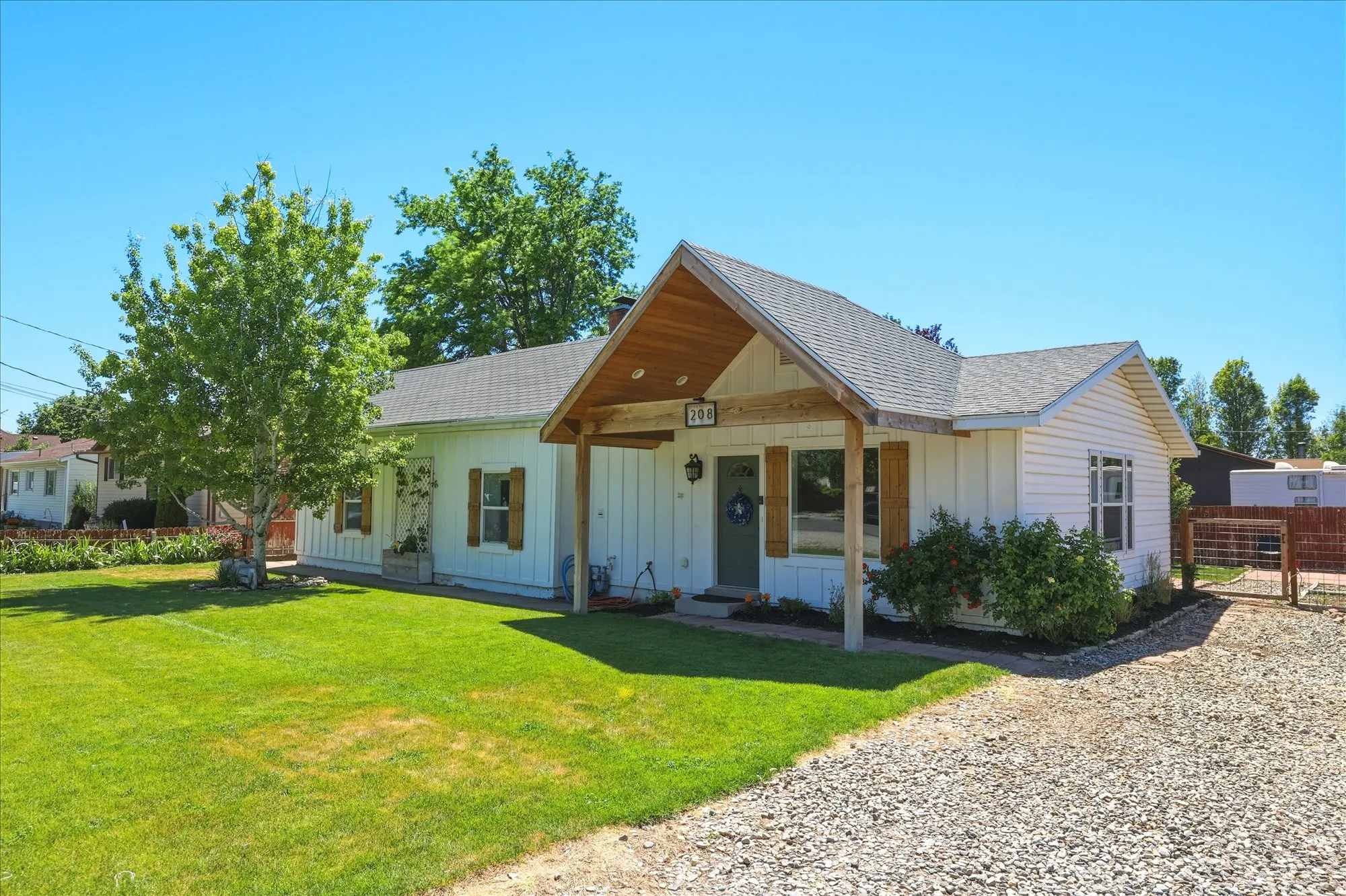 White siding exterior with a wood-accented covered entry, featuring a gravel driveway, manicured lawn, and mature tree landscaping