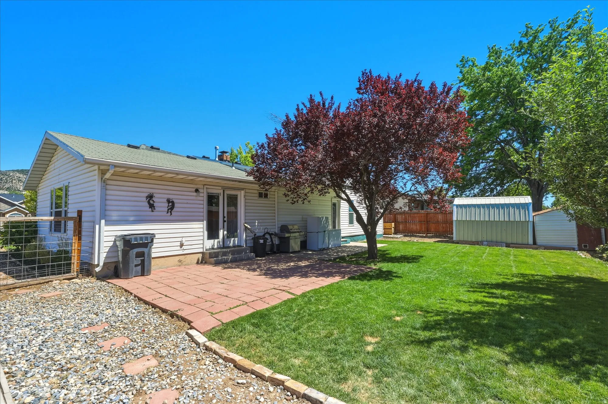 Expansive backyard featuring a red paver patio, a vibrant red-leafed tree, a large metal storage shed, a smaller white storage shed, and a privacy fence