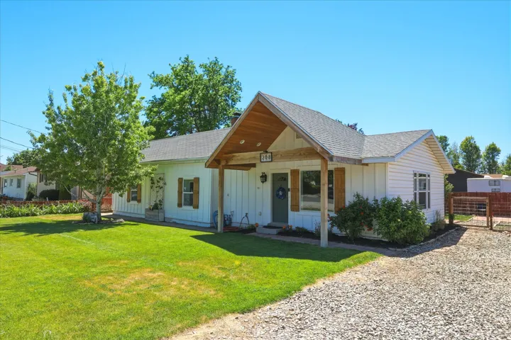White siding exterior with a wood-accented covered entry, featuring a gravel driveway, manicured lawn, and mature tree landscaping