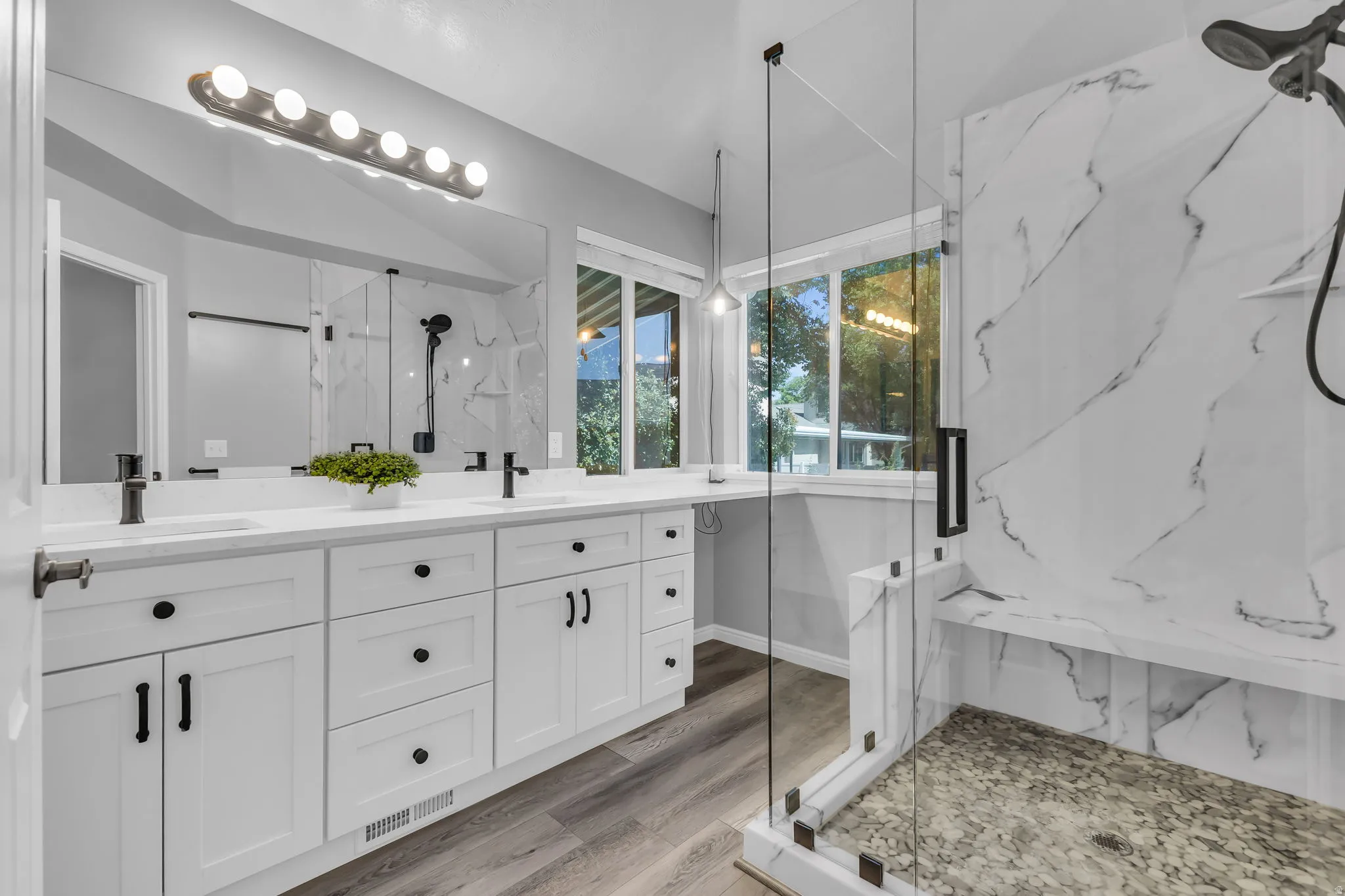 Walk-in shower with clear glass enclosure and stone base, dual vanity with undermount sinks, matte black fixtures, wood-finish flooring, and white cabinetry