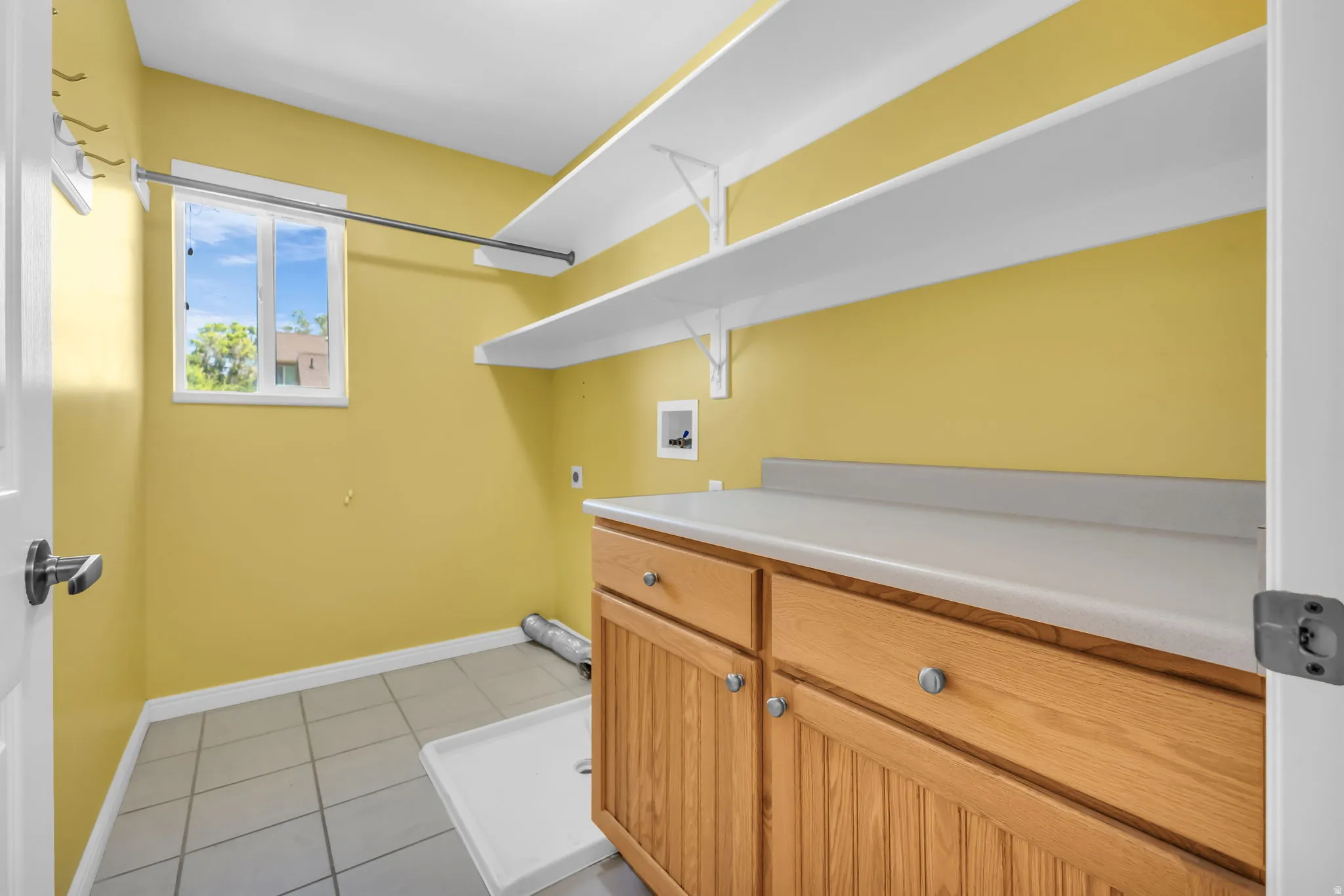 Laundry area featuring a window, tile flooring, built-in shelving, a clothes hanging rod, and a wood-finish cabinet with a countertop