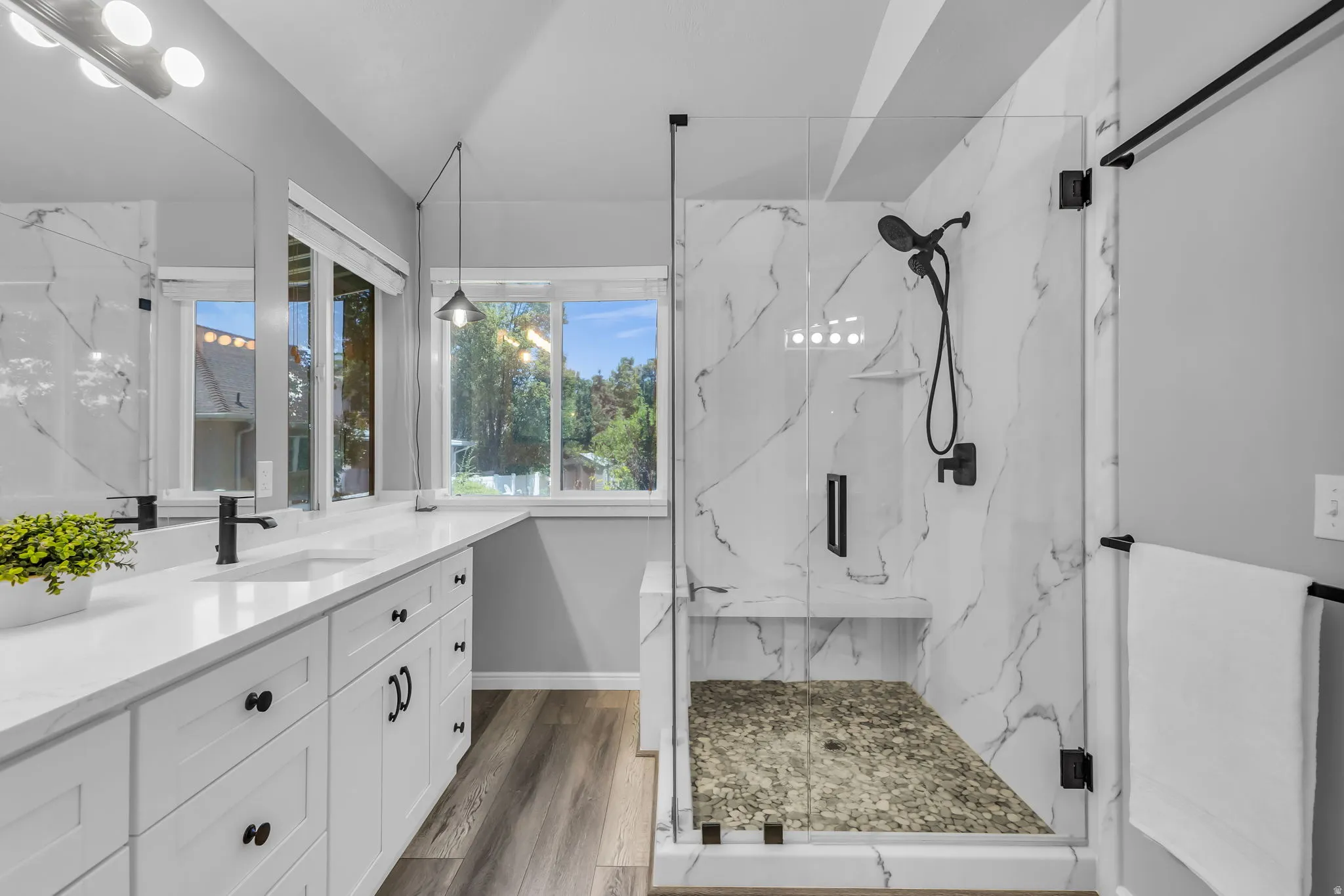 Walk-in shower with clear glass enclosure and stone base, dual vanity with undermount sinks, matte black fixtures, wood-finish flooring, and white cabinetry