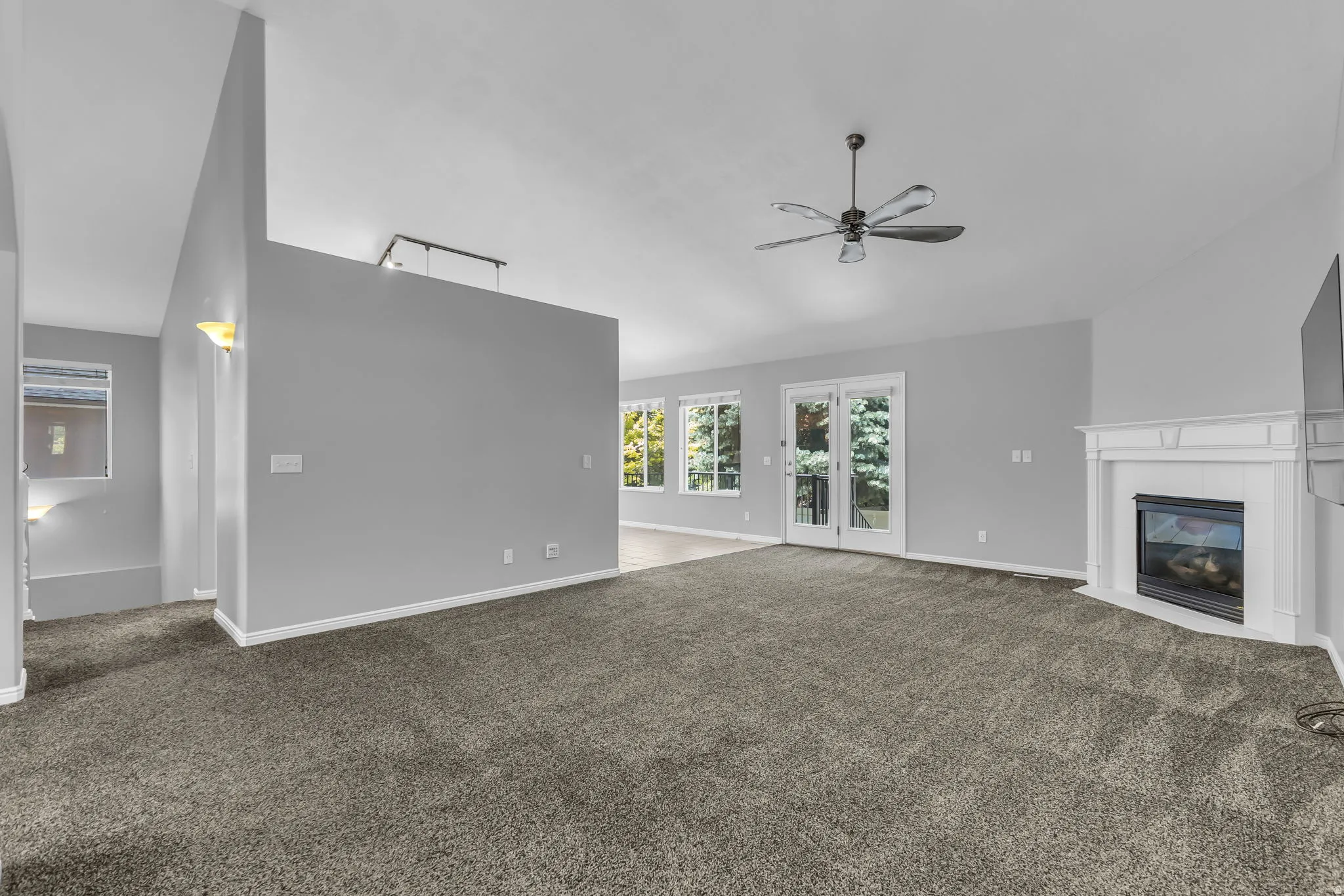 Spacious living area featuring vaulted ceilings, a ceiling fan, a corner fireplace with a white mantel, and double French doors opening to the deck.