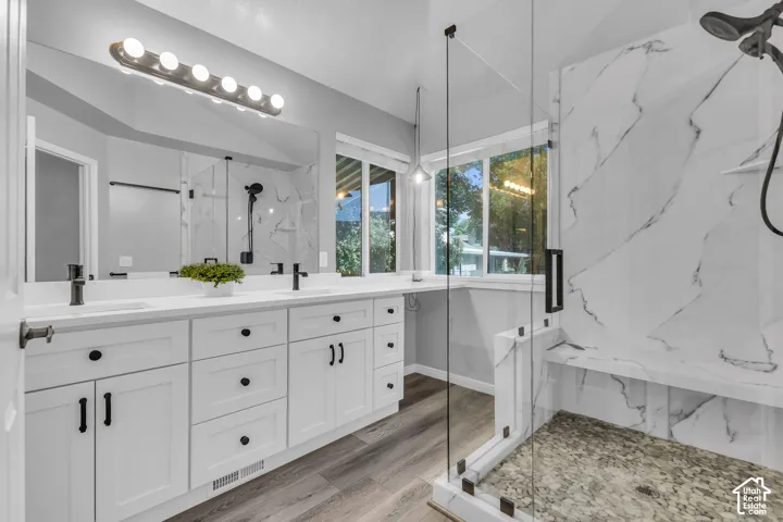 Walk-in shower with clear glass enclosure and stone base, dual vanity with undermount sinks, matte black fixtures, wood-finish flooring, and white cabinetry
