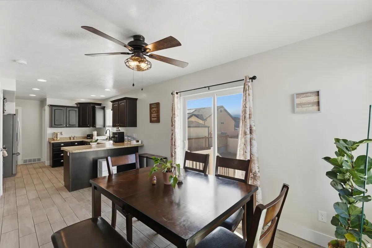 Open-concept dining and kitchen area featuring wood-finish flooring, dark wood cabinetry, light-toned countertops, a ceiling fan with integrated lighting, and a sliding glass door