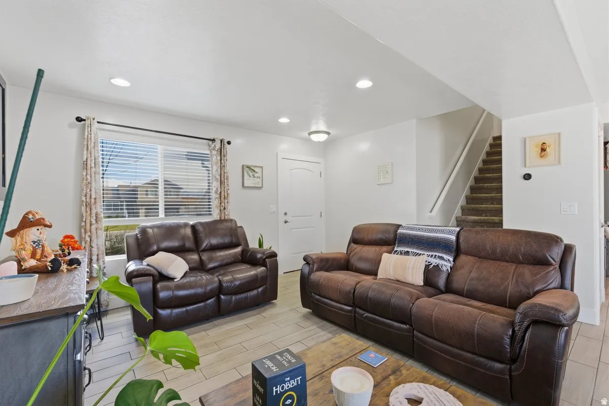 Living area featuring light wood-finish flooring, recessed lighting, a front-facing window with blinds, and a staircase with carpeted treads
