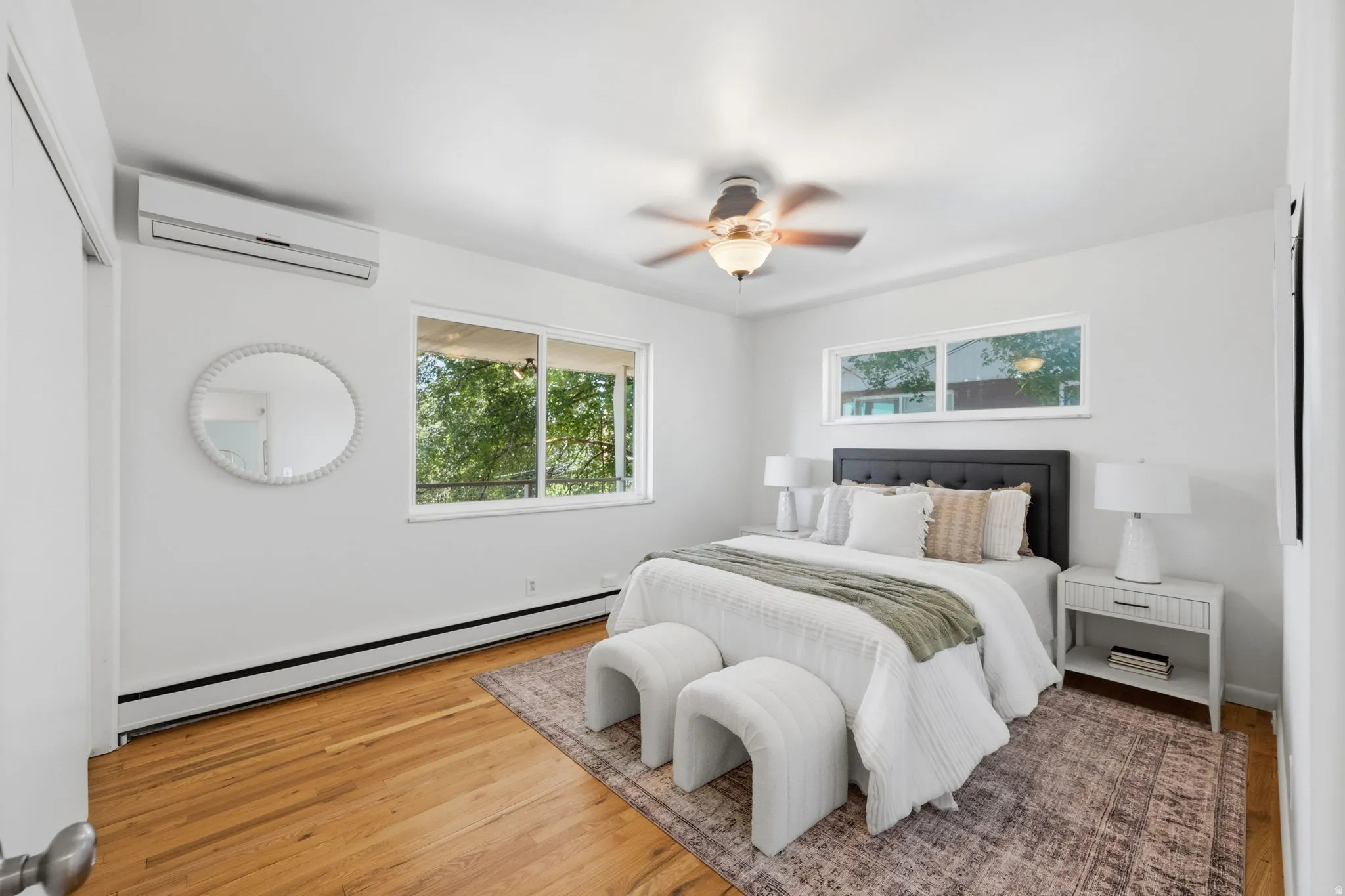 Bright room featuring two windows, wood-finish flooring, a ceiling fan with integrated lighting, and a wall-mounted climate control unit