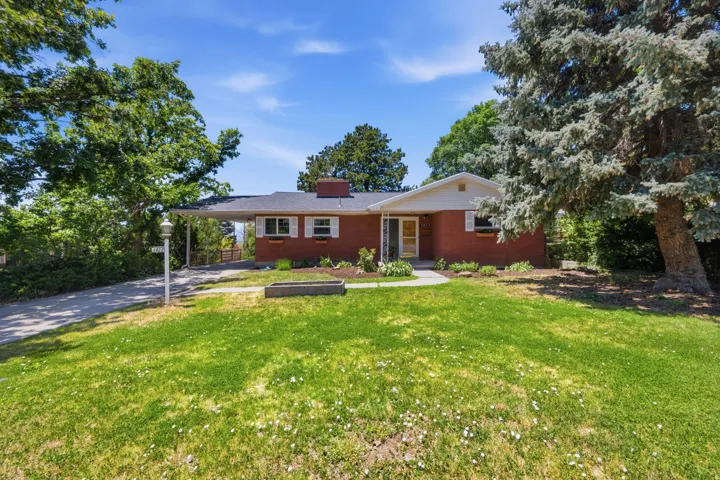 Red brick exterior residence featuring a front lawn with established trees, a curved concrete pathway, a covered carport, and a prominent chimney