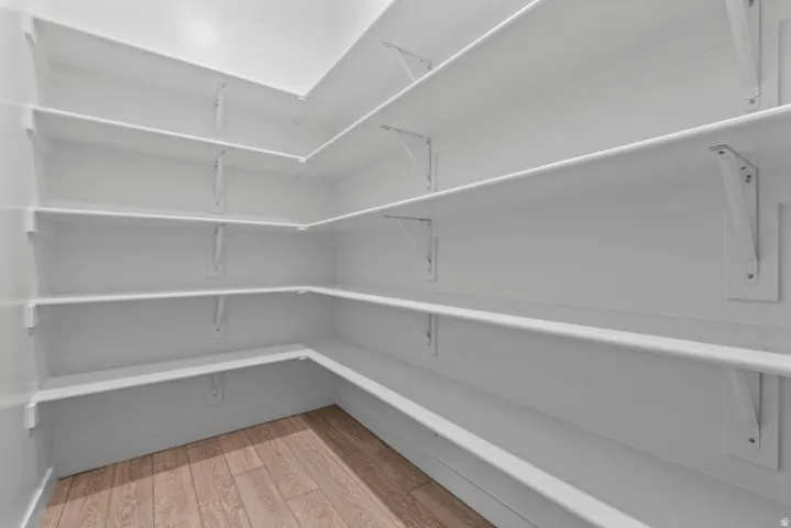 Walk-in pantry featuring extensive white shelving on multiple walls, solid wood-finish flooring, and bright overhead lighting