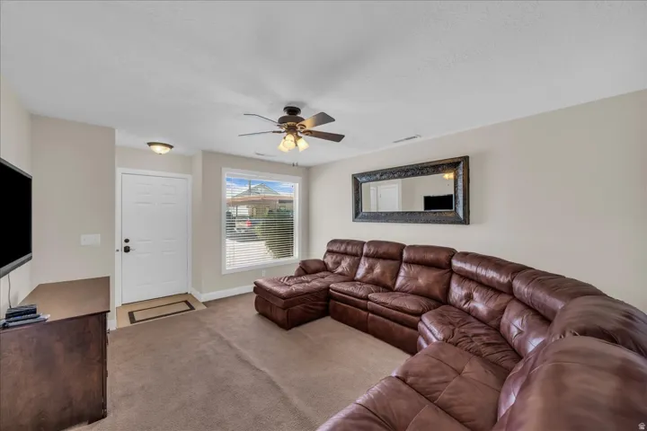 Inviting living area featuring a large window, neutral wall tones, textured carpeting, a ceiling fan with integrated lighting, and a white paneled entry door