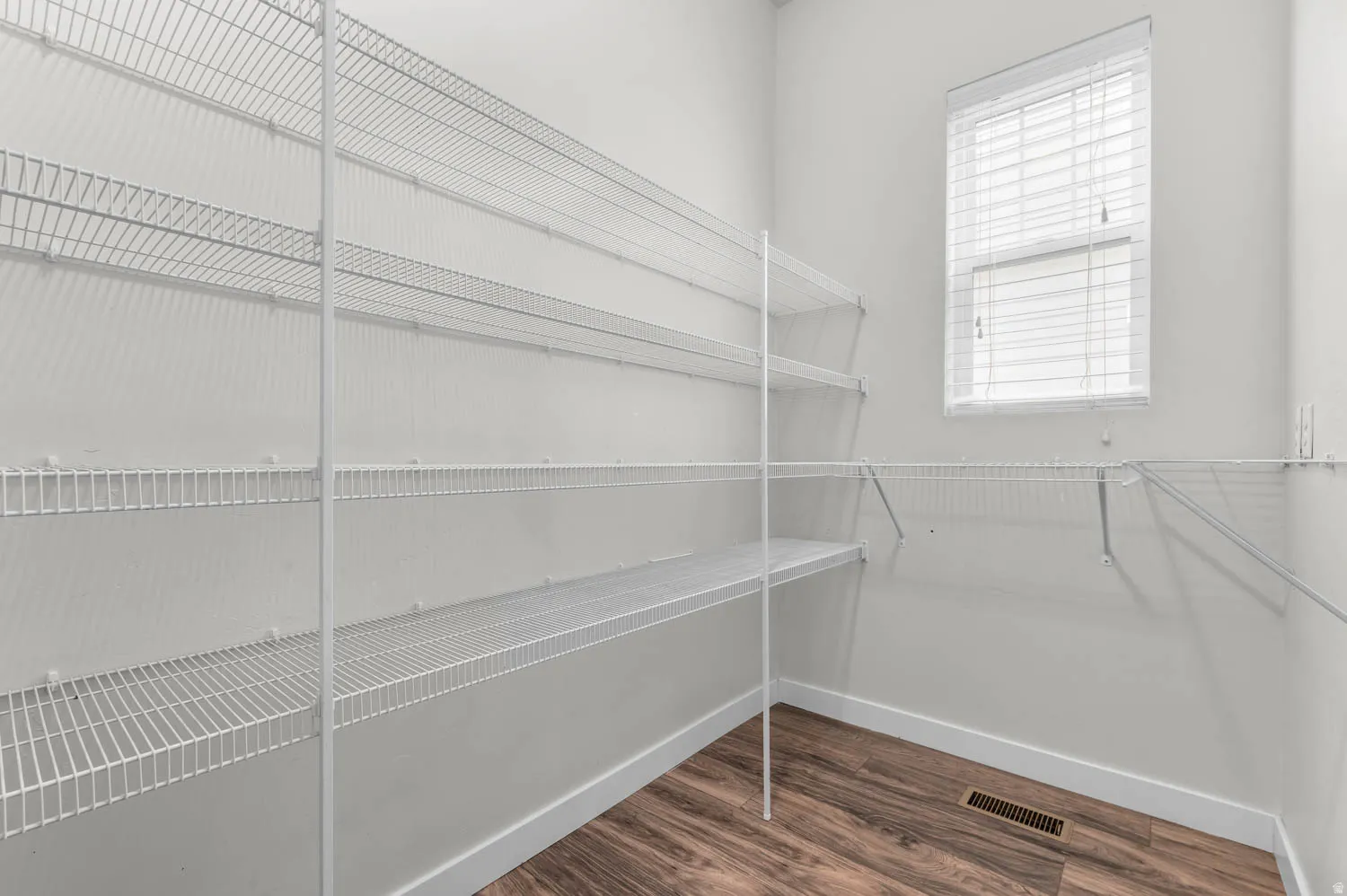 Spacious storage area featuring extensive wire shelving, natural light from a window with blinds, wood-finish flooring, and crisp white baseboards