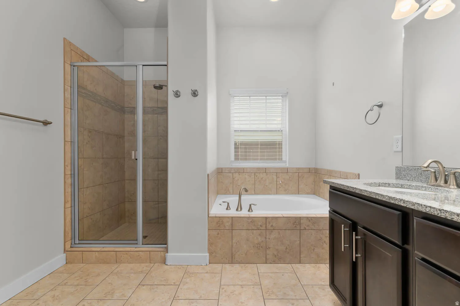 Bathroom featuring a tiled soaking tub, an enclosed shower with a glass door, a double vanity with stone countertops, and beige tile flooring