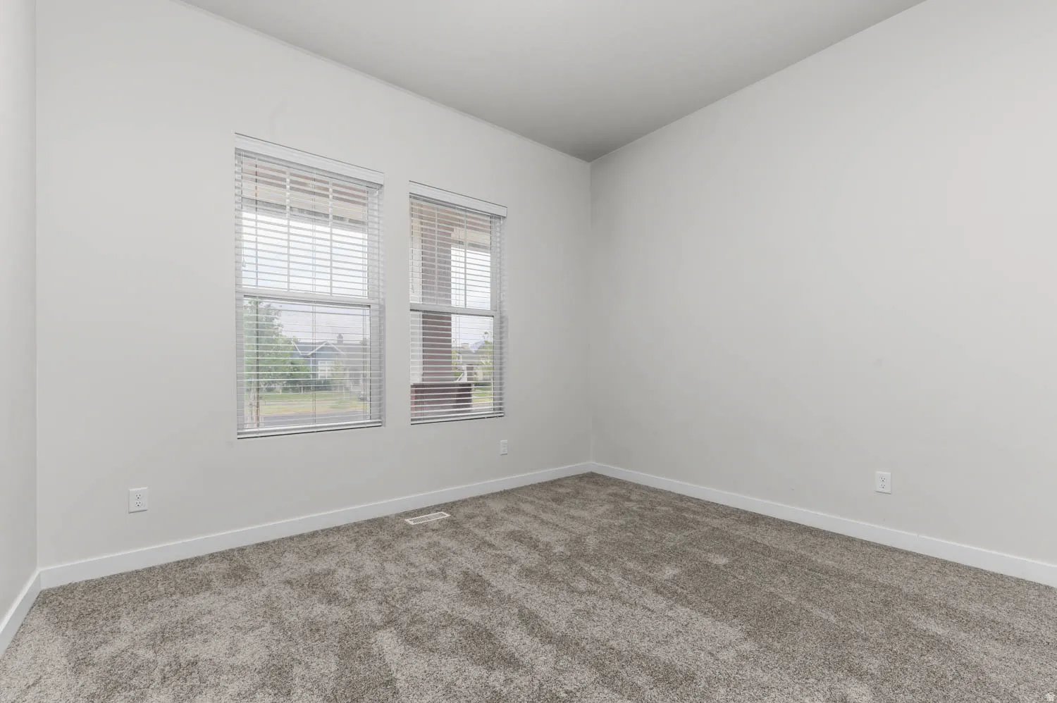 Room with two windows featuring horizontal blinds, light gray textured carpeting, white baseboards, and neutral wall paint