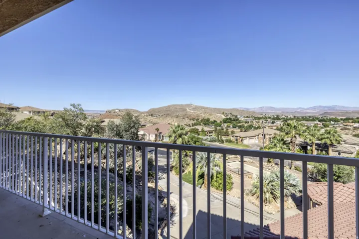 Balcony with a white metal railing overlooking a residential area with varied rooflines and extensive landscaping, framed by distant hills