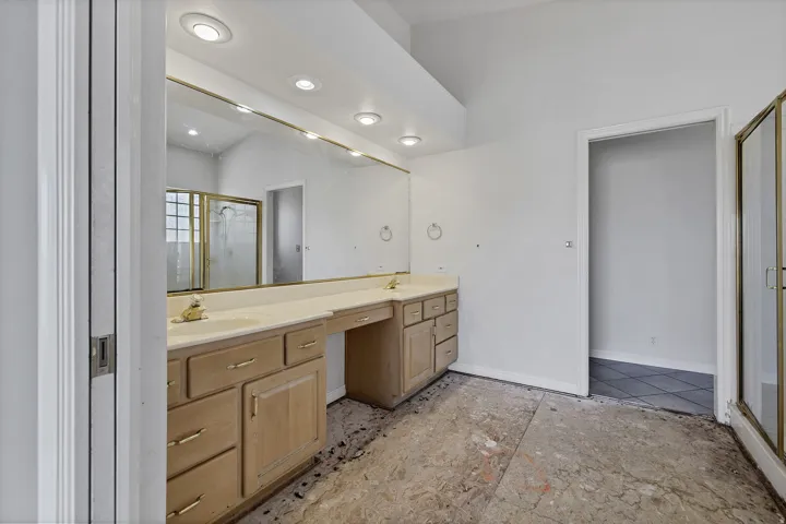 Vaulted ceiling bathroom featuring a dual vanity with light-tone cabinetry, a gold-framed wall mirror with integrated lighting, and a glass-enclosed shower with gold-tone framing