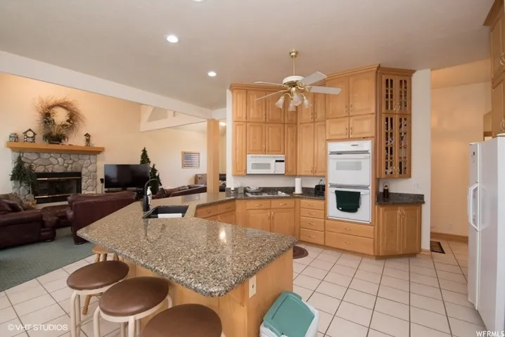 Expansive kitchen featuring a large island with a dark countertop and sink, light wood cabinetry, white tile flooring, and integrated white appliances