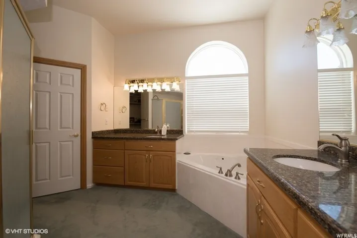 Bathroom featuring a built-in jetted tub, two vanity cabinets with integrated sinks, granite-finish countertops, and arched windows with blinds