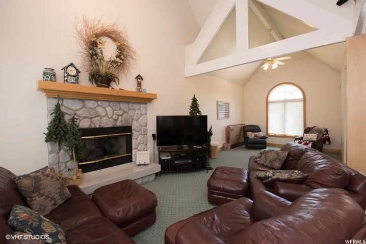 Spacious living area featuring a stone fireplace with a wood mantle, vaulted ceilings with exposed beams, a ceiling fan, and an arched window