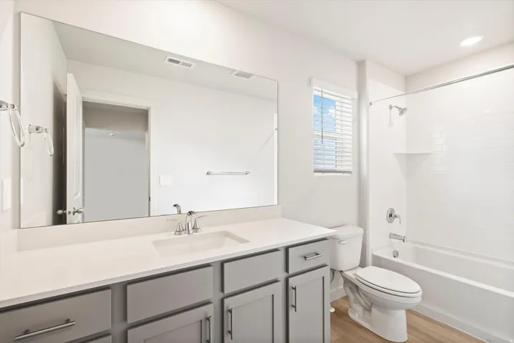 **DISLCAIMER** Interior package may vary. Bathroom featuring a light gray vanity with a white countertop and integrated sink, a large frameless mirror, a white bathtub with subway tile surround, and wood-finish flooring