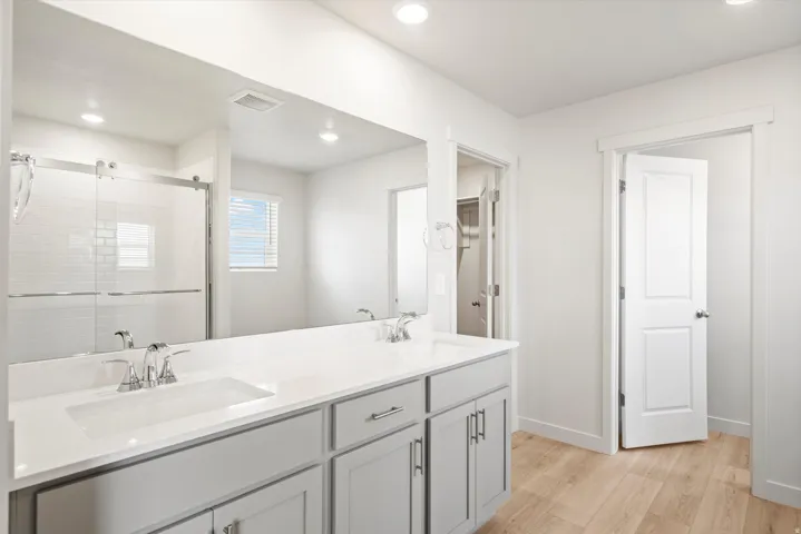 **DISLCAIMER** Interior package may vary. Bathroom featuring a dual-sink vanity with light gray cabinetry and chrome hardware, a full-width mirror, wood-finish flooring, and a glass-enclosed shower with white subway tile