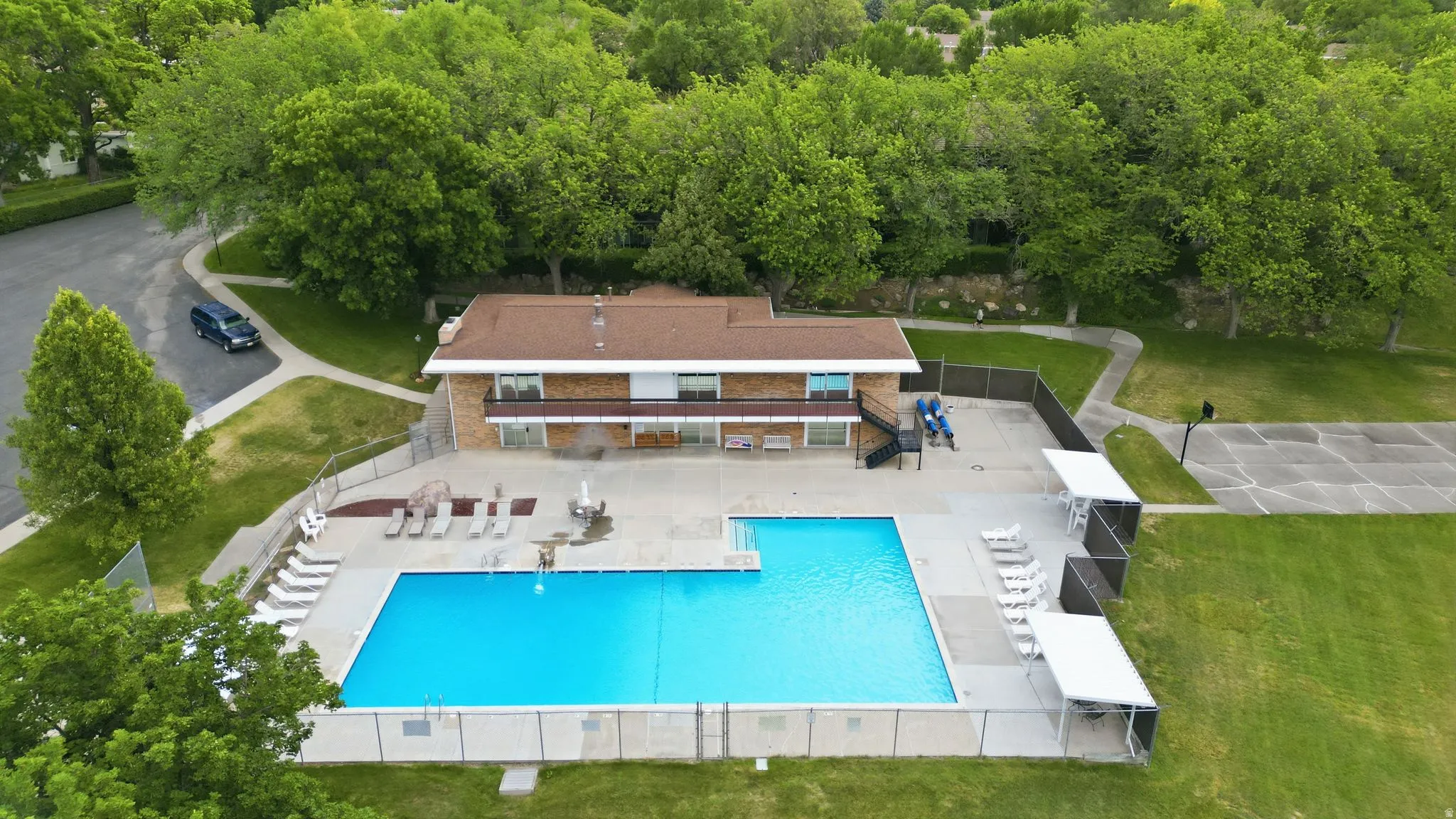 Fenced outdoor swimming pool with surrounding concrete decking, lounge chairs, and an adjacent two-story brick building with large grass fields