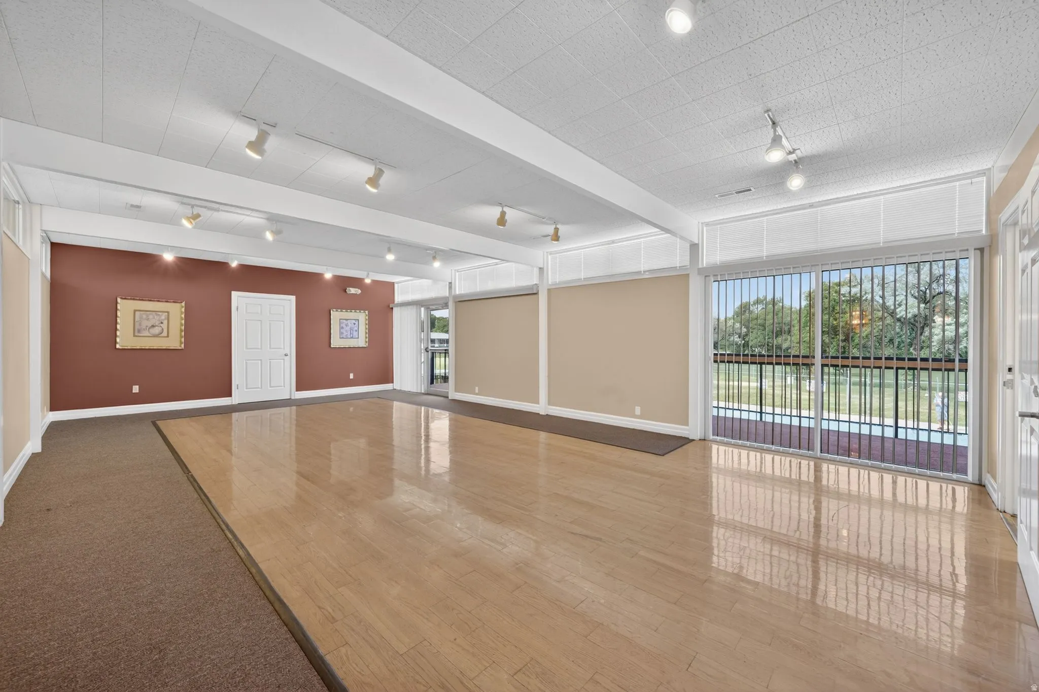 Club house spacious hosting room with wood-finish flooring, track lighting, a drop ceiling, and a sliding glass door leading to a balcony with a metal railing