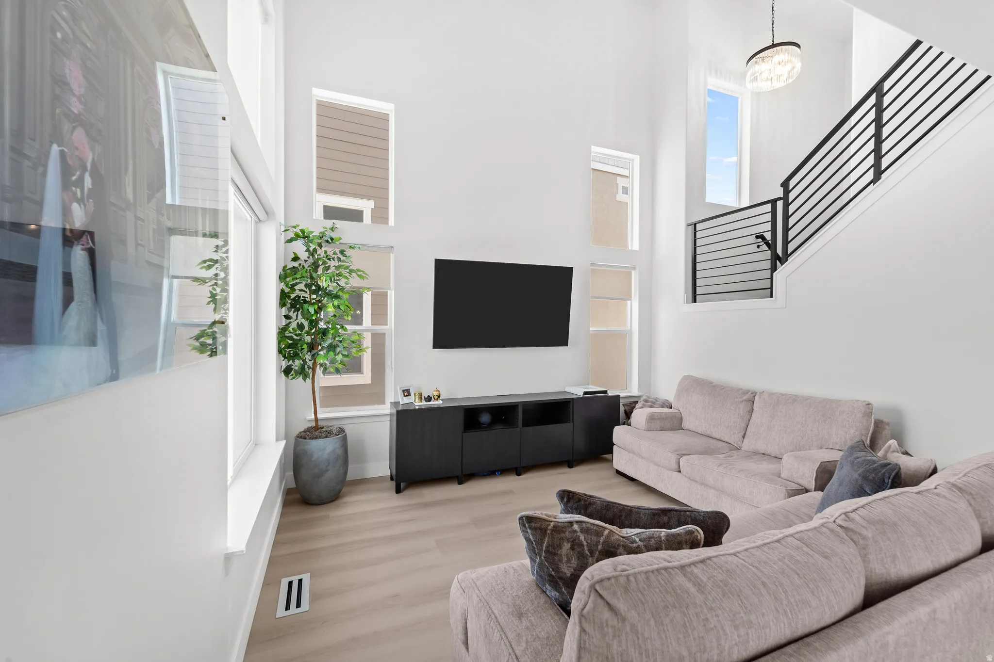 Vaulted living space featuring an open-riser staircase with black metal balusters, high ceilings, light wood-finish flooring, a wall-mounted media console, and a contemporary chandelier