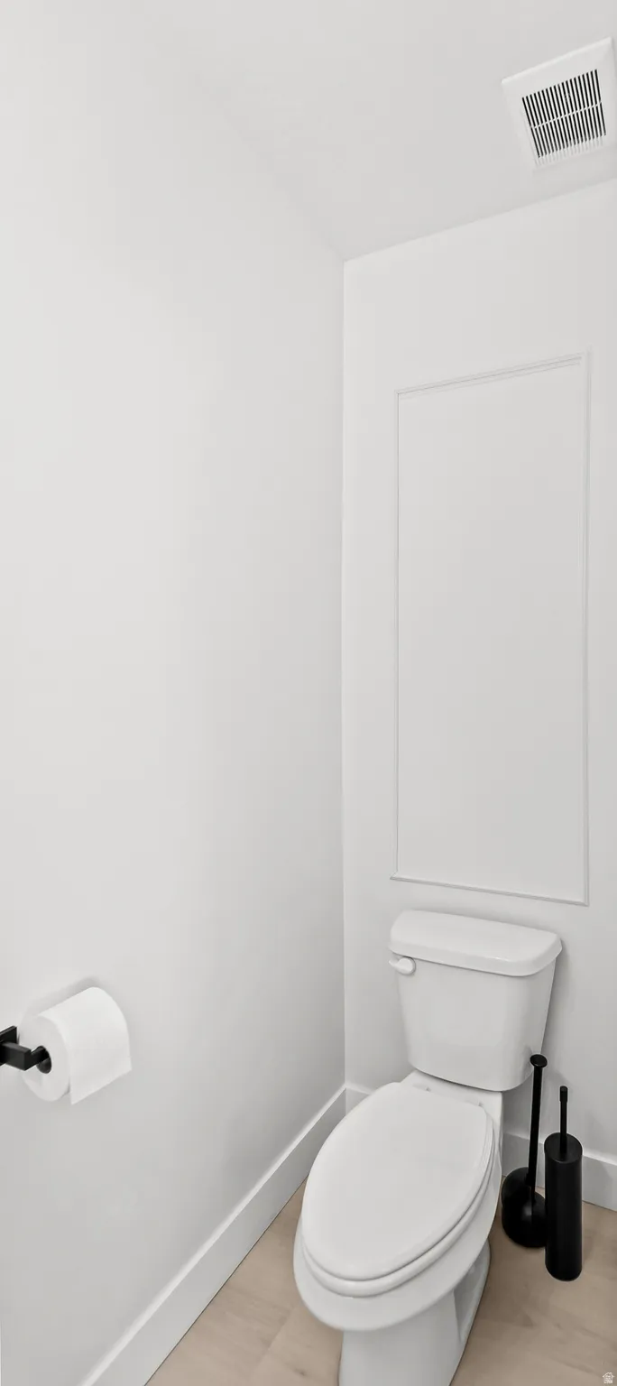 White toilet with a built-in panel accent wall, light wood-finish flooring, and a black wall-mounted toilet paper holder