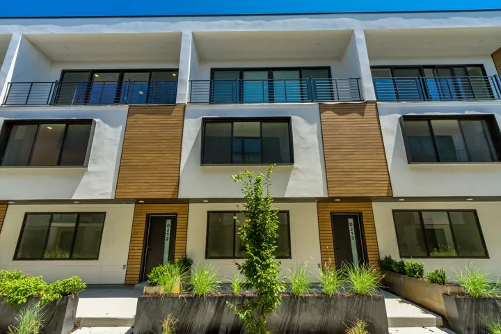 Contemporary multi-level facade featuring white stucco, wood-finish paneling, multiple balconies with black railings, large dark-framed windows, and a dark entry door