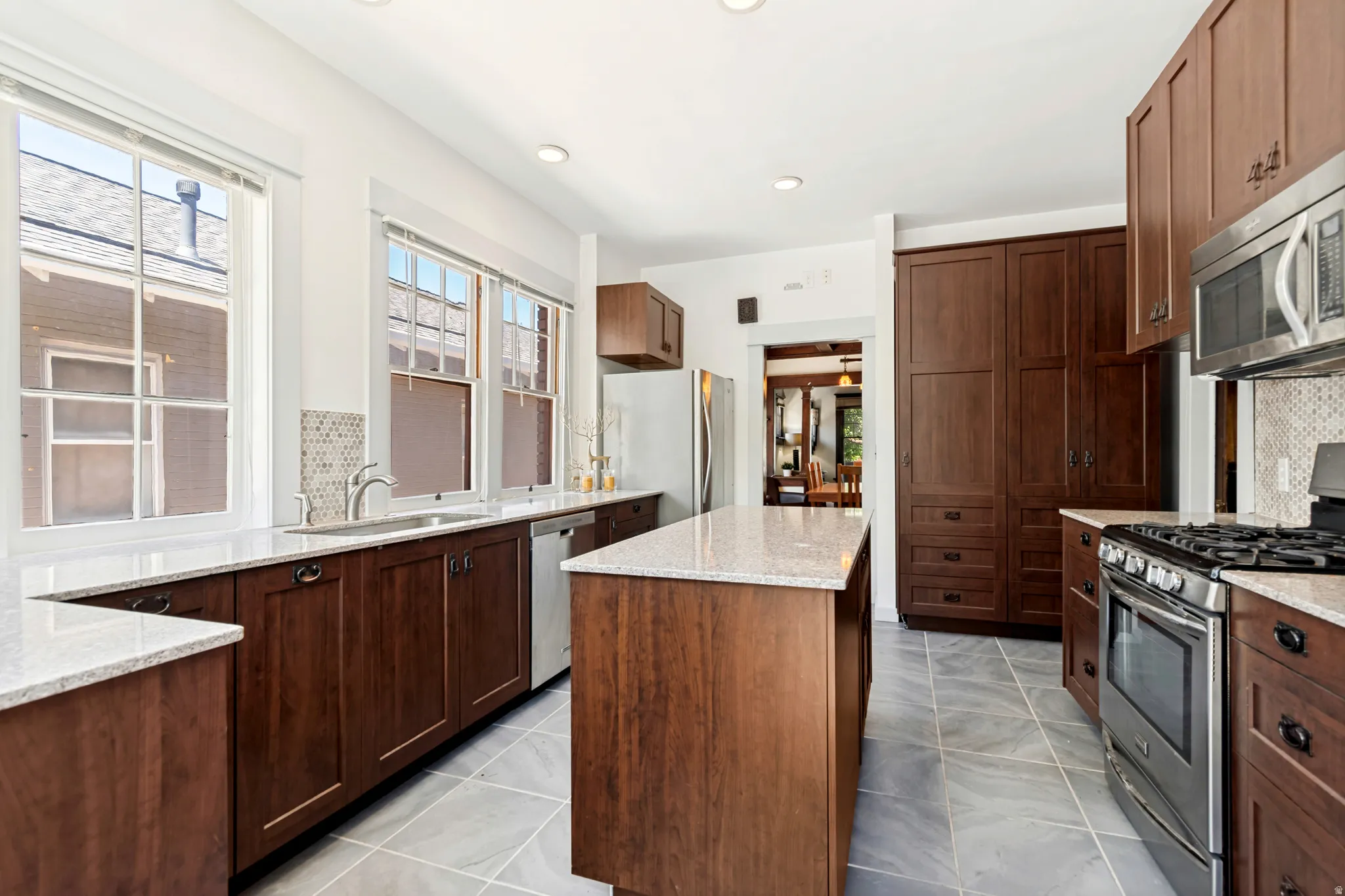 Kitchen featuring wood-finish cabinetry, light-toned countertops, a central island, stainless steel appliances, and a mosaic tile backsplash