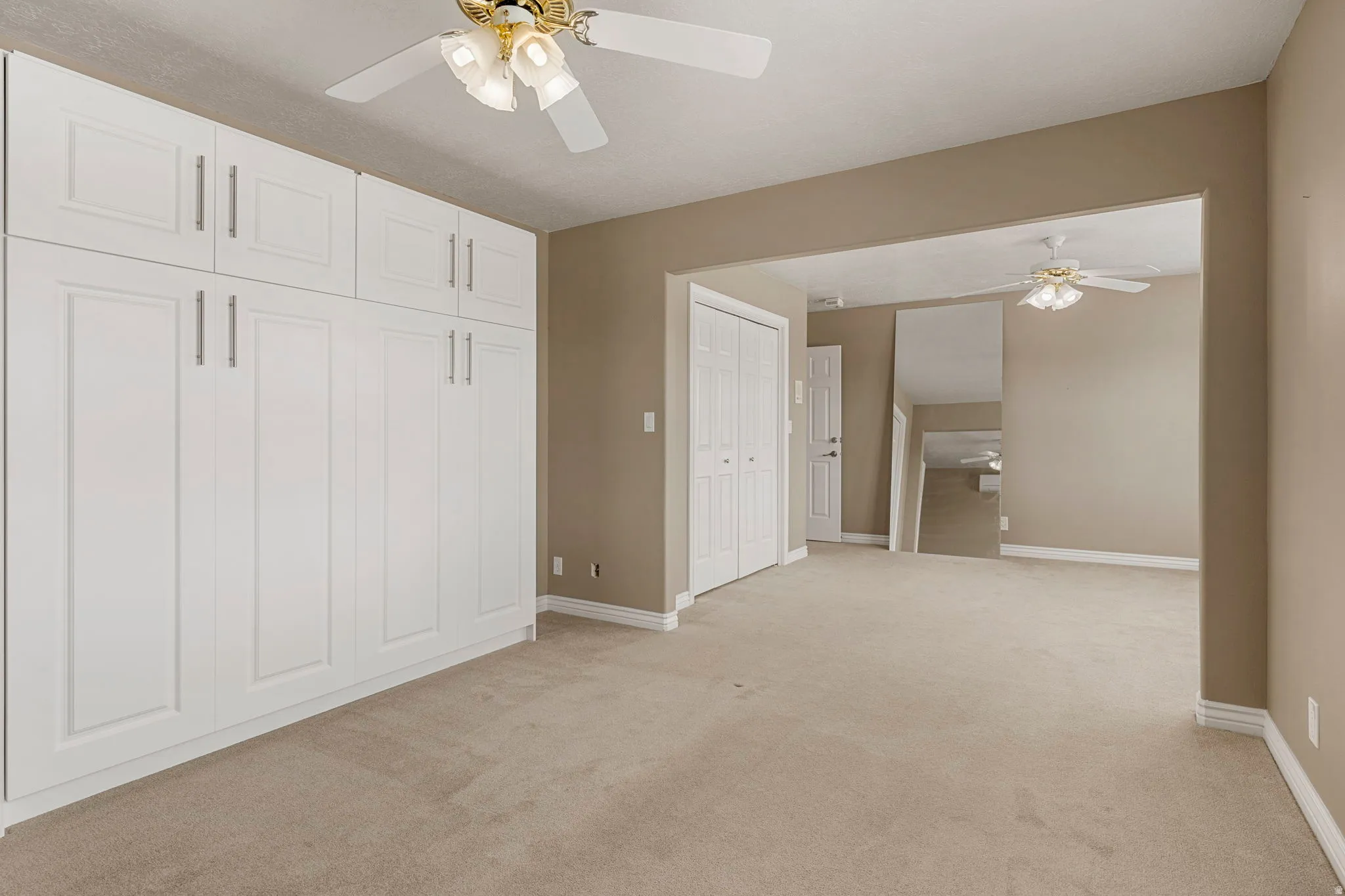 Bedroom featuring extensive built-in white panel cabinetry with silver-tone hardware, plush beige carpeting, a ceiling fan with integrated lighting, and neutral wall tones