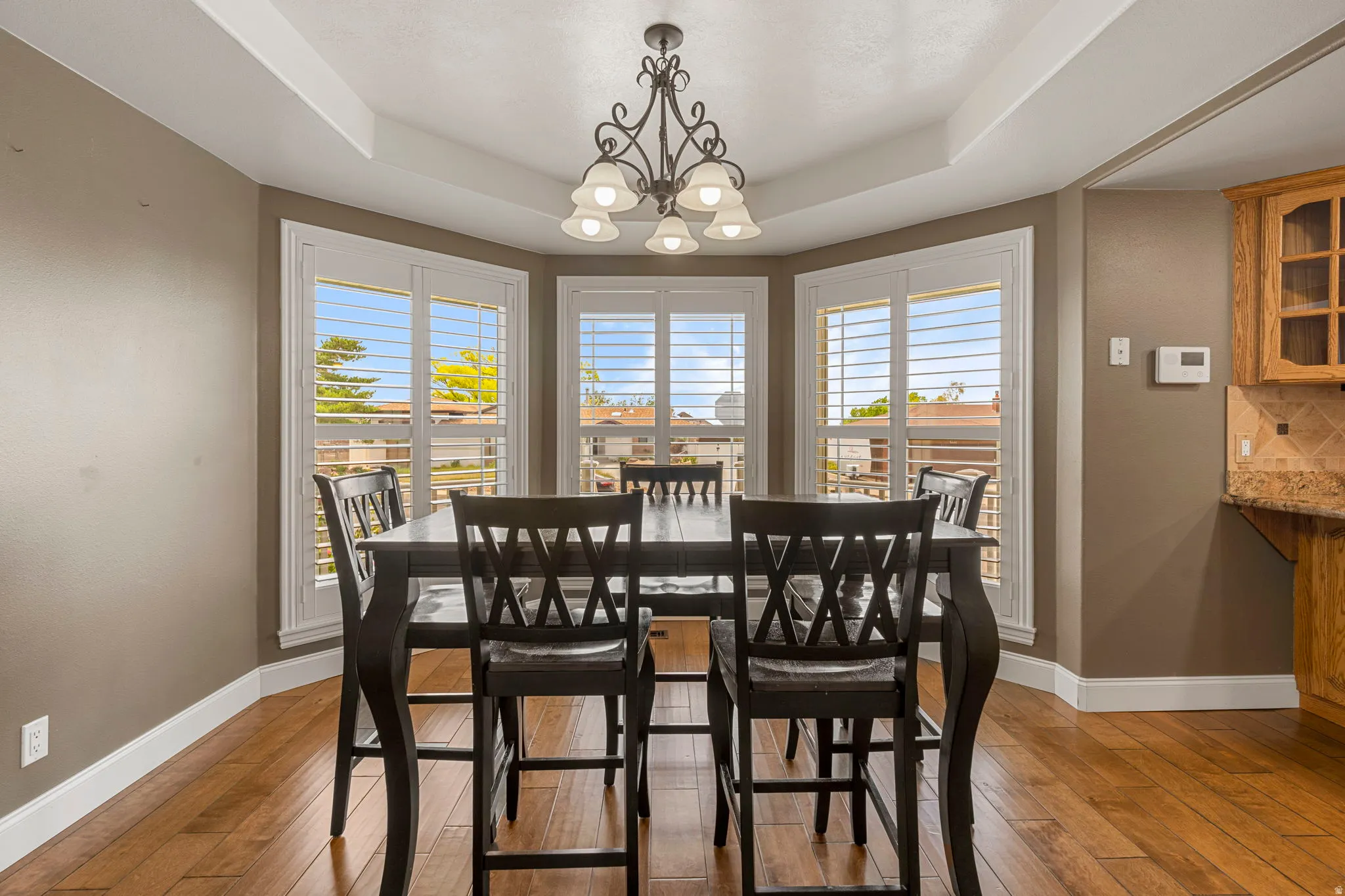 Dining area featuring a tray ceiling with a decorative chandelier, multiple bay windows with white plantation shutters, and wood-finish flooring