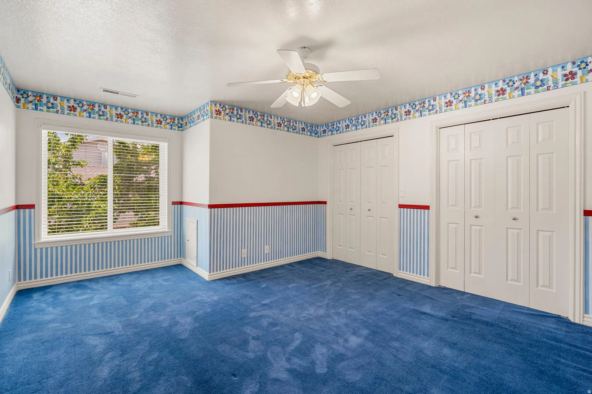Spacious room featuring plush blue carpeting, a large window with horizontal blinds, and a ceiling fan with integrated lighting