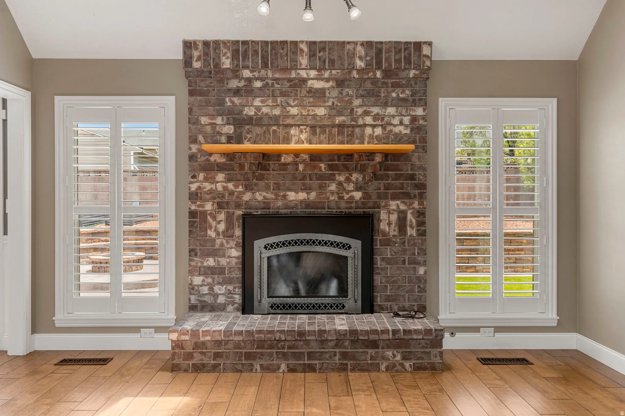 Brick fireplace with a wood mantel, flanked by two windows with white plantation shutters, set against neutral-toned walls and wood-finish flooring