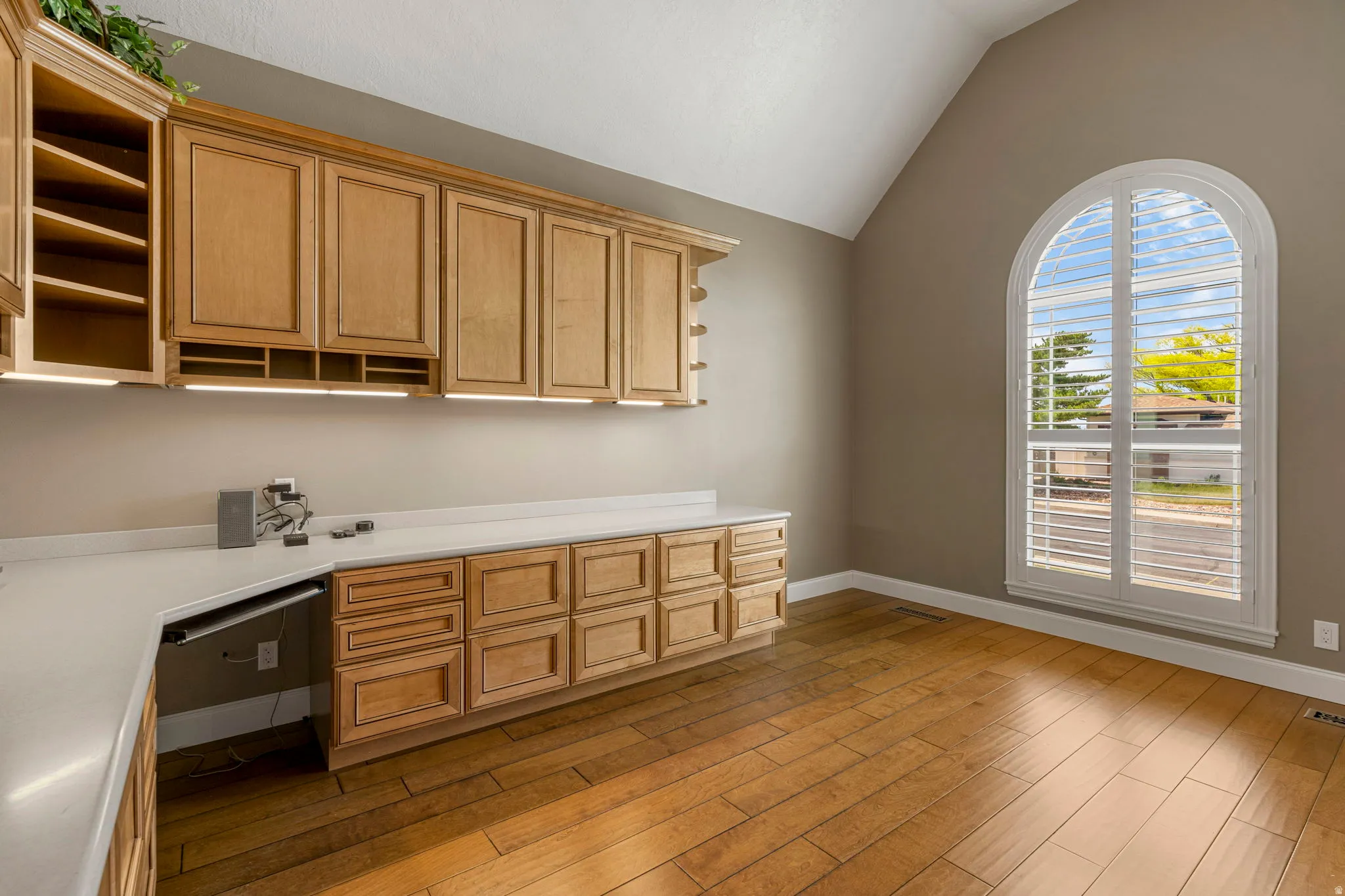 Arch-top window with plantation shutters, vaulted ceiling, extensive built-in cabinetry, under-cabinet lighting, and wood-finish flooring