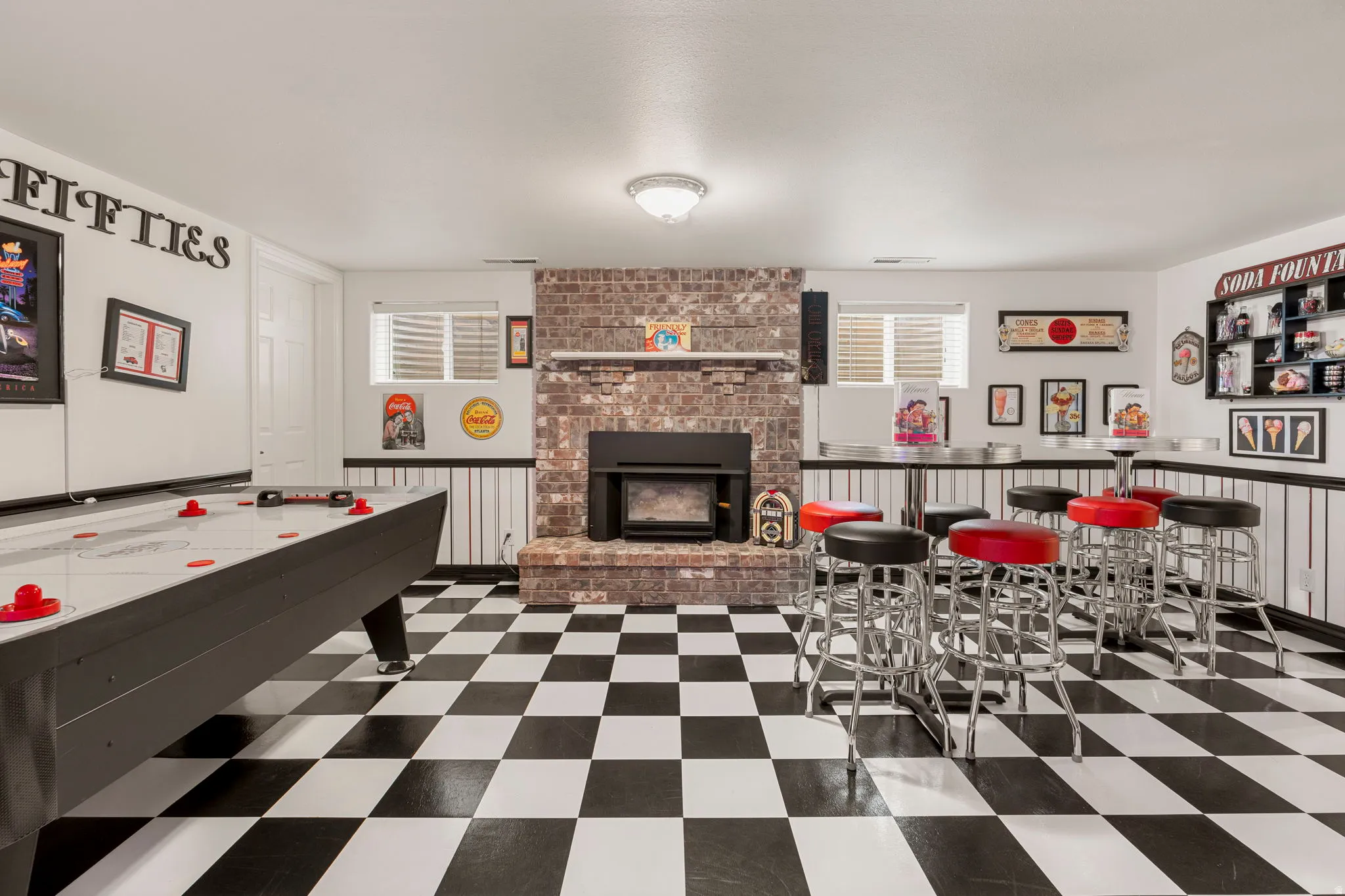 Spacious recreational area featuring checkerboard flooring, a brick-clad fireplace with a mantel, white wainscoting, and recessed windows with blinds