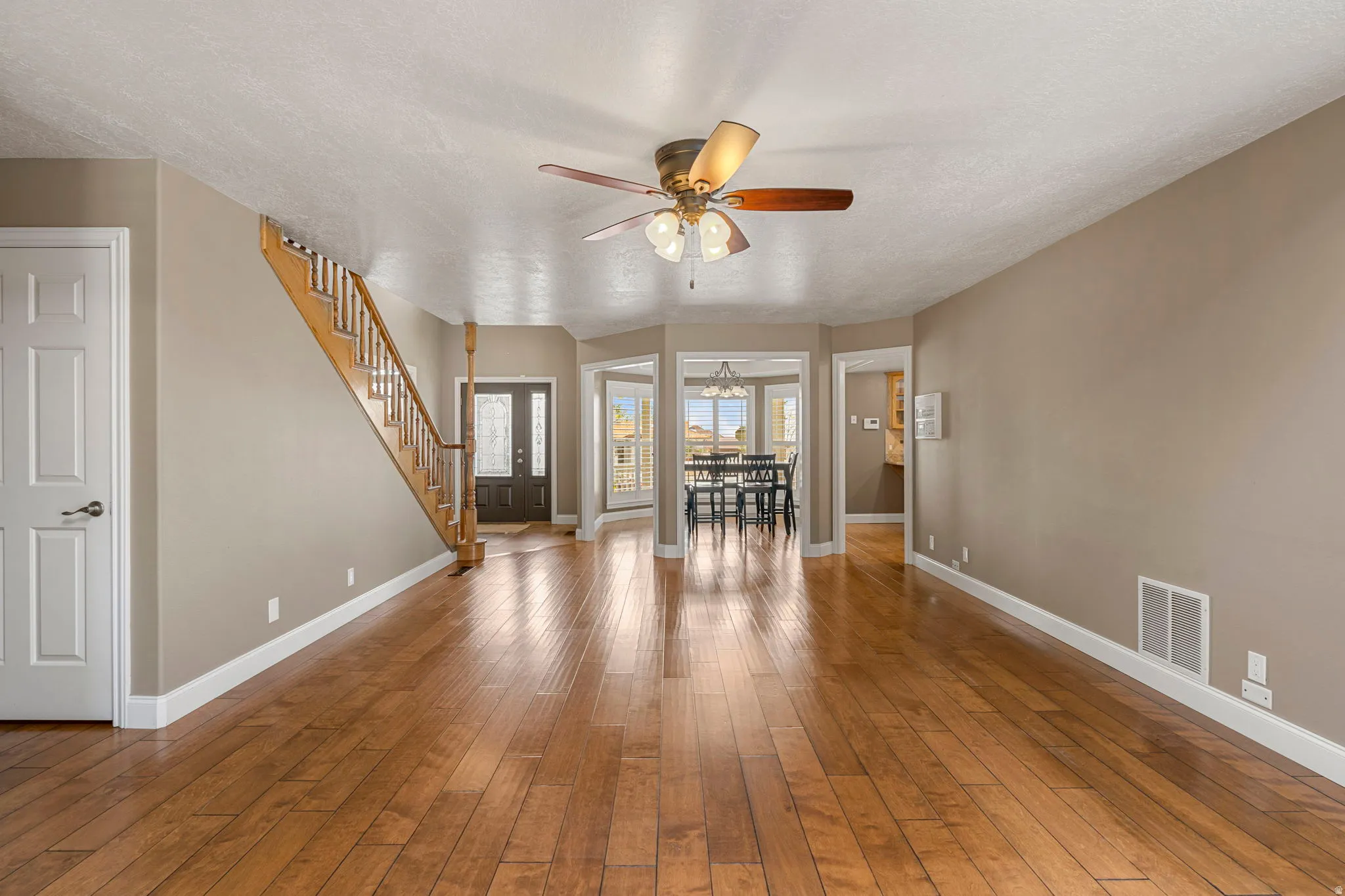Spacious living area featuring wood-finish flooring, a ceiling fan, and access to an entryway with a dark wood door and staircase with wood railing
