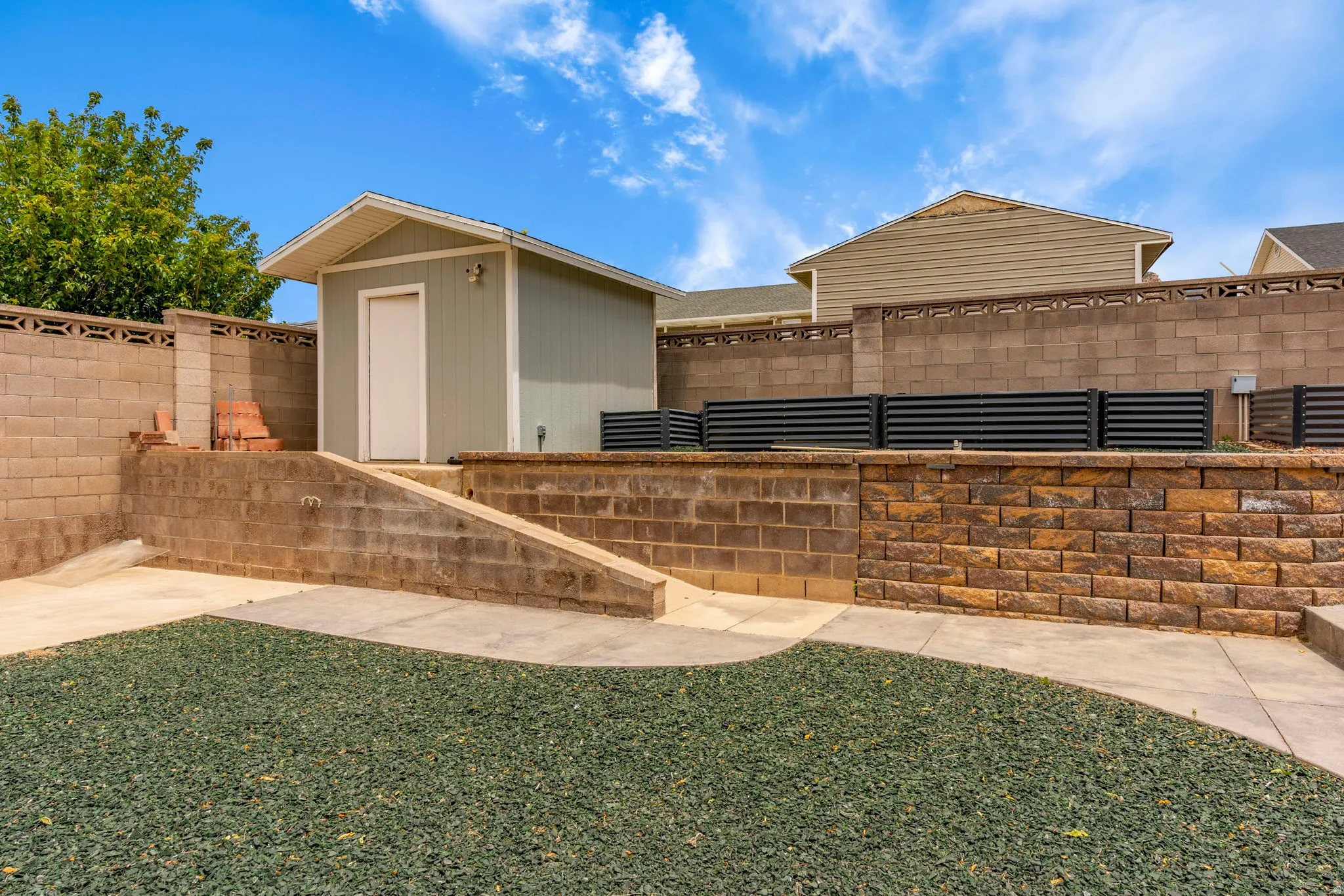 Terraced backyard featuring a dedicated storage shed with a gable roof, concrete block retaining walls, a decorative stone veneer retaining wall, a paved concrete patio, and a ground cover planting bed