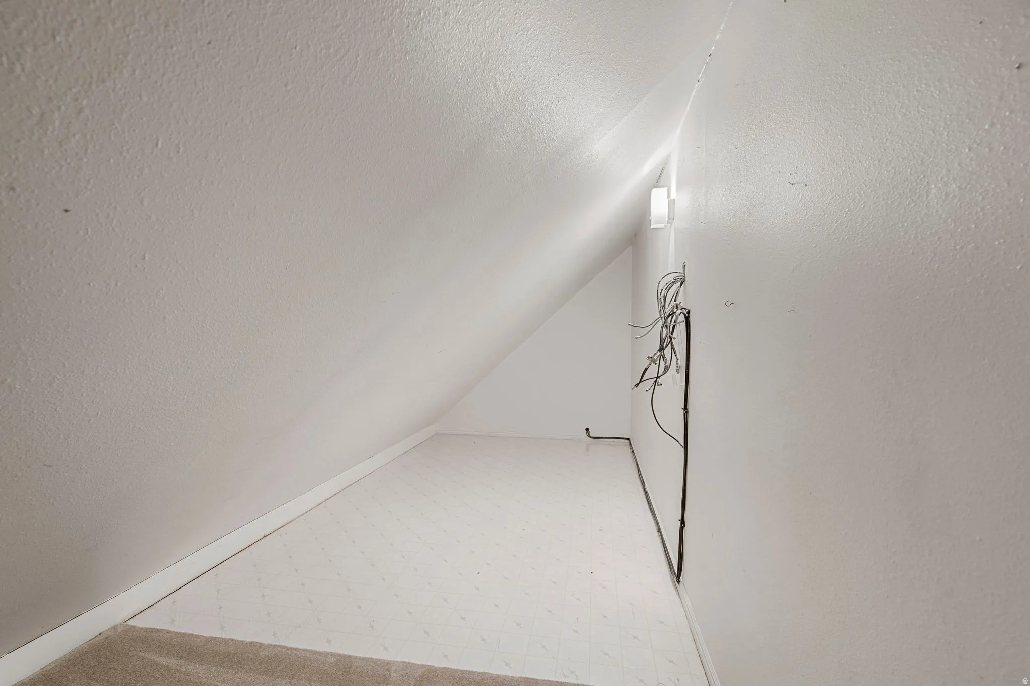 Sloping ceiling dormer space with white wall paint, wall-mounted light fixture, and a mix of carpet and textured flooring