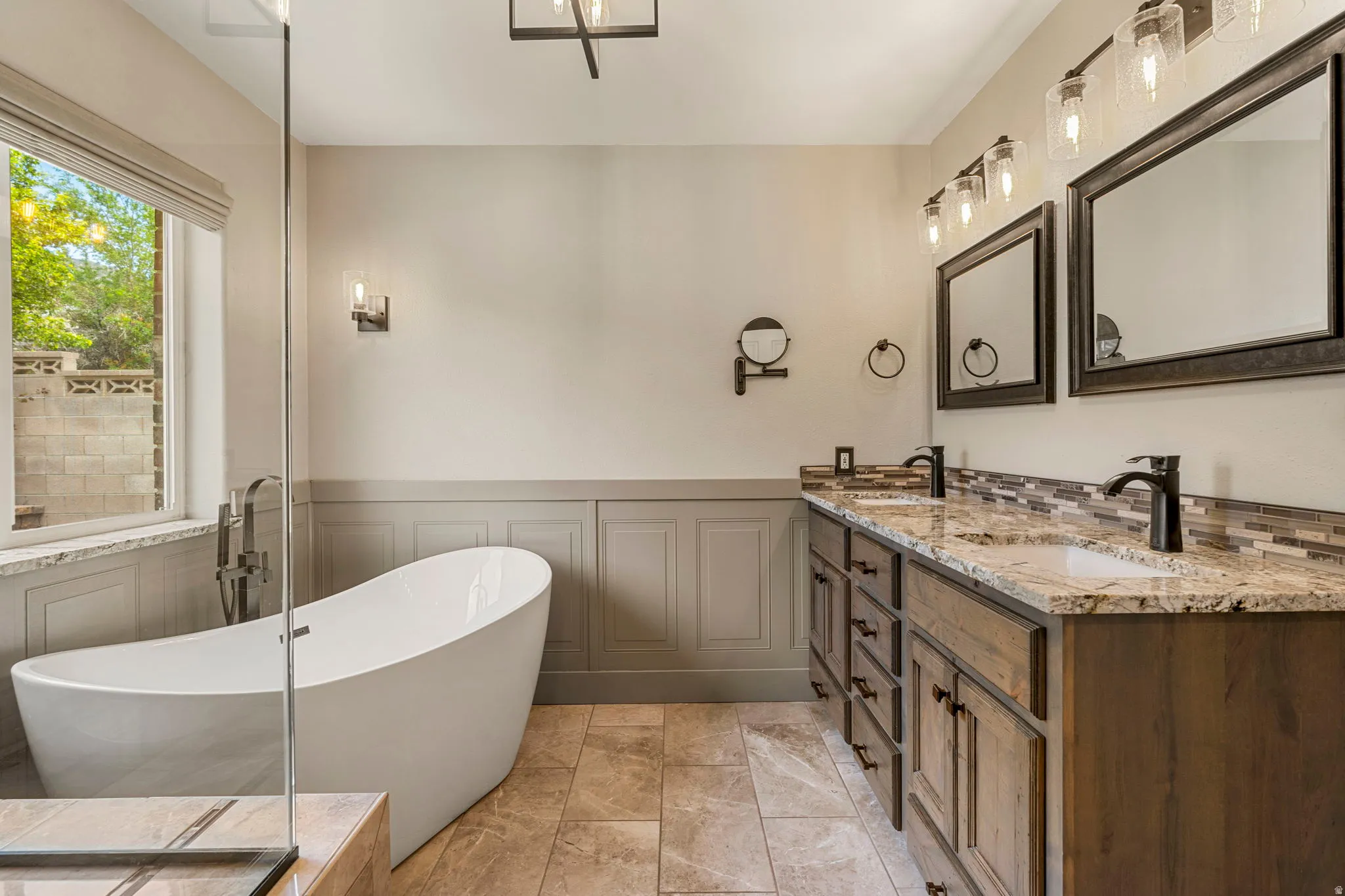 Freestanding soaking tub with a floor-mounted faucet, dual wood-finish vanity with stone countertops and undermount sinks, tile backsplash, and tile flooring