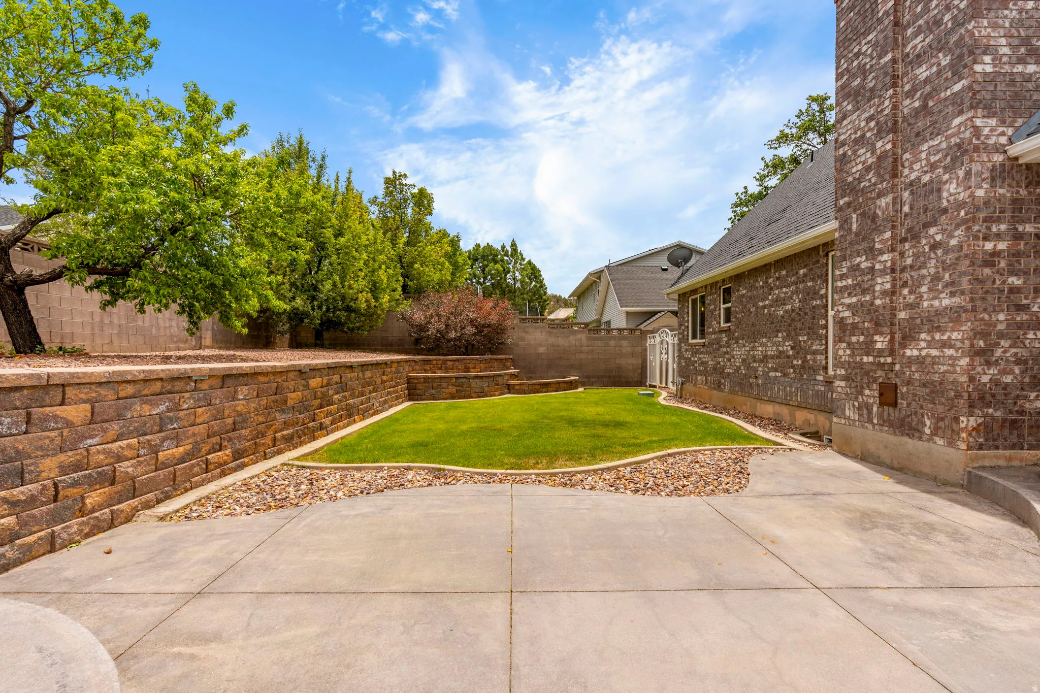 Expansive concrete patio area with a manicured lawn, tiered retaining walls constructed from stone, and mature landscaping
