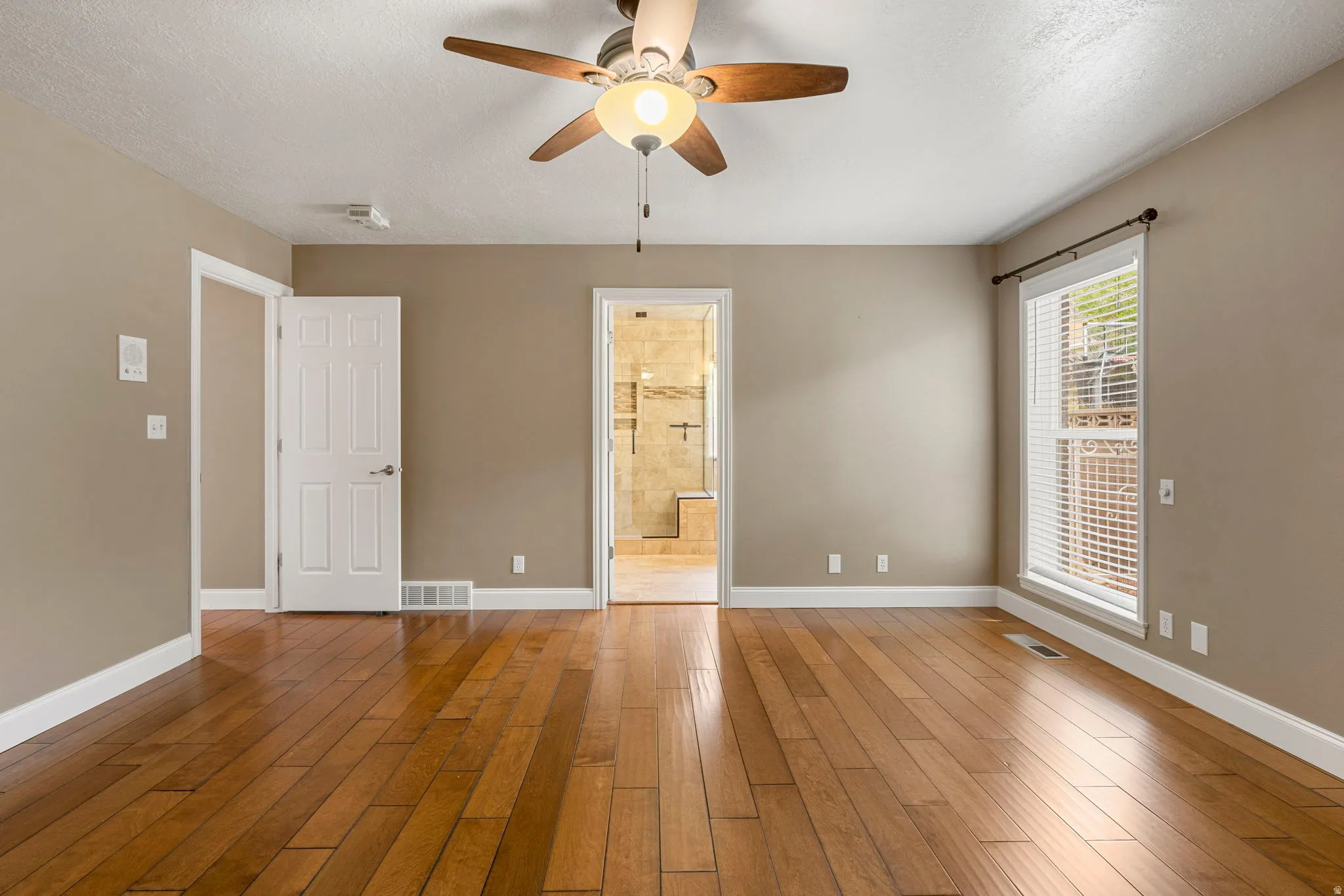 Spacious room featuring wood-finish flooring, a ceiling fan, and ample natural light from a large window with blinds