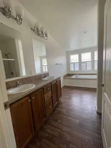 Dual vanity with wood-finish cabinetry, built-in bathtub, wood-finish flooring, and window shutters