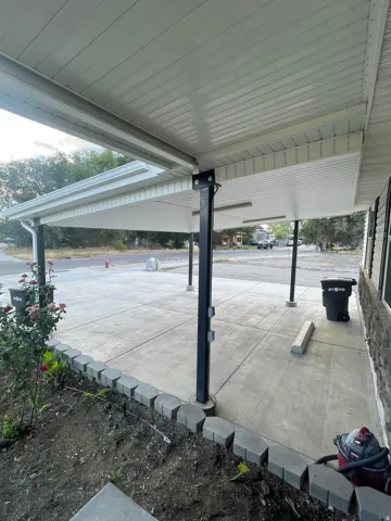 Covered concrete patio with recessed lighting, supported by black structural pillars, bordered by a decorative stone landscape edge