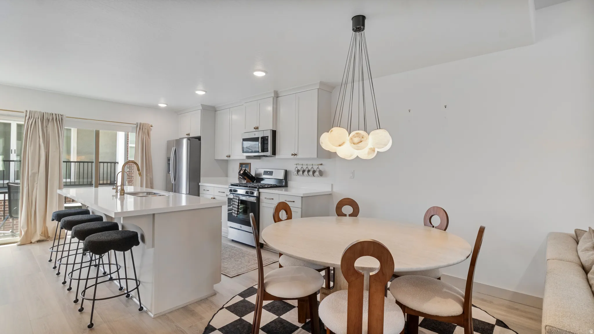Open-concept kitchen and dining area featuring light wood-finish flooring, a kitchen island with seating, white shaker cabinetry, stainless steel appliances, and a contemporary dining room chandelier