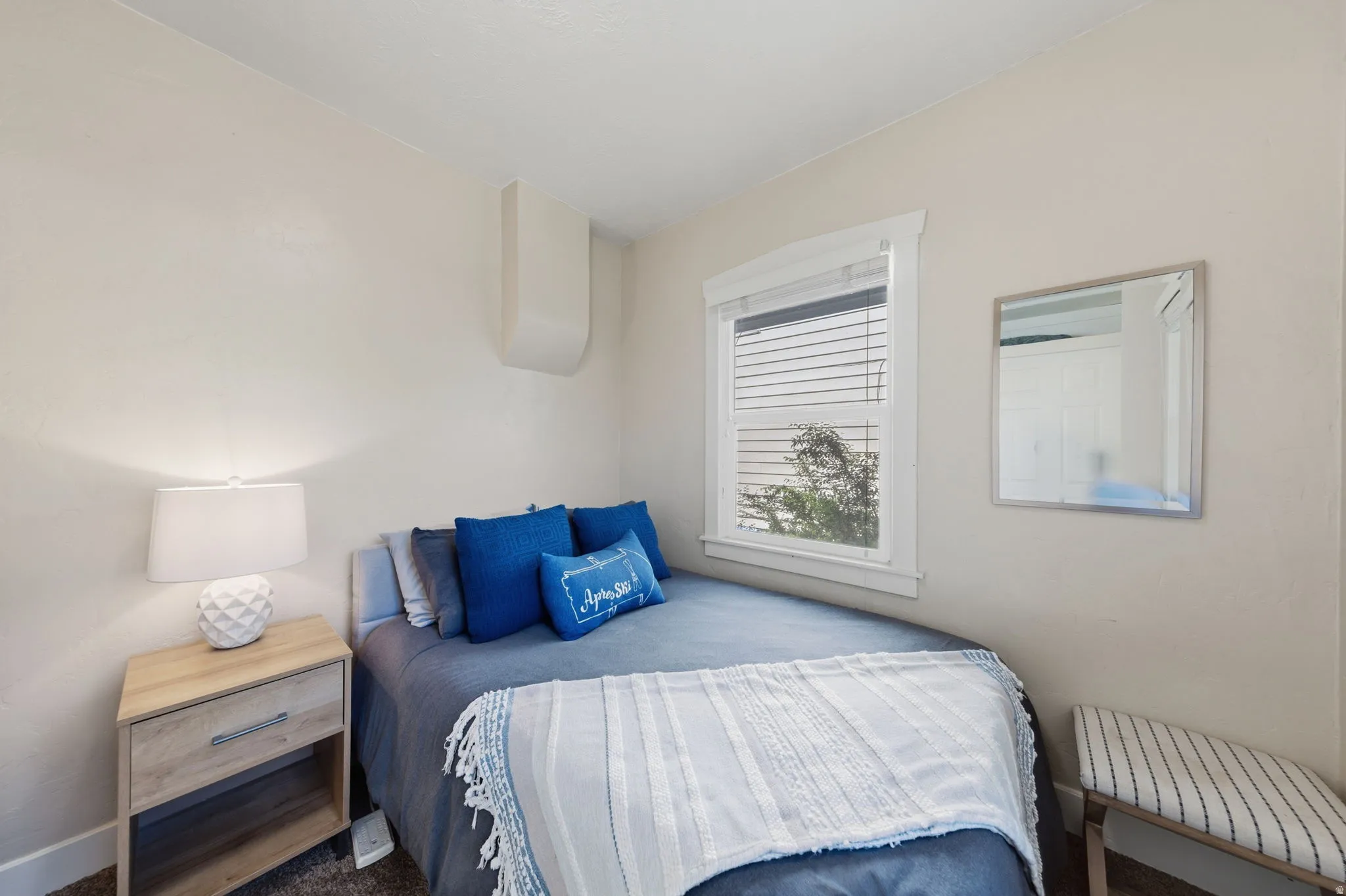 Bright room featuring a single window with white blinds, neutral wall tones, wood-finish flooring, and a wall-mounted mirror
