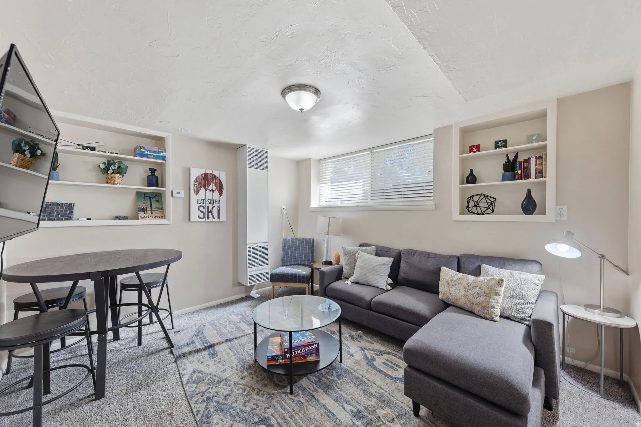 Carpeted living area featuring recessed shelving, baseboard heating, a ceiling-mounted light fixture, and horizontal blinds