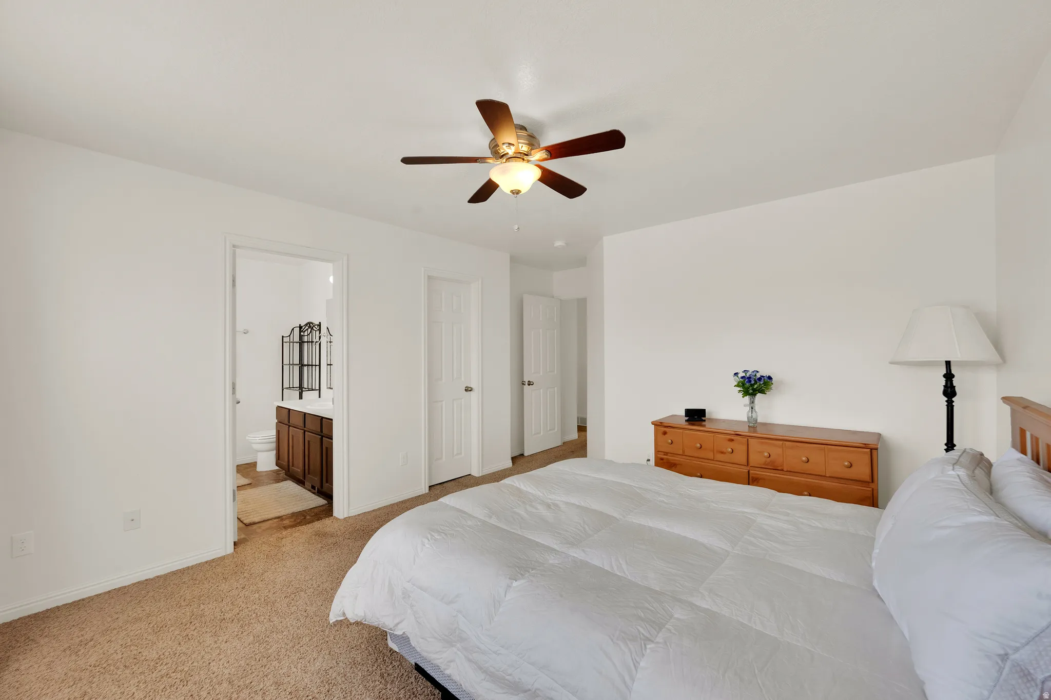 Carpeted bedroom with a ceiling fan, featuring a doorway to a bathroom with a dark wood-finish vanity and white toilet