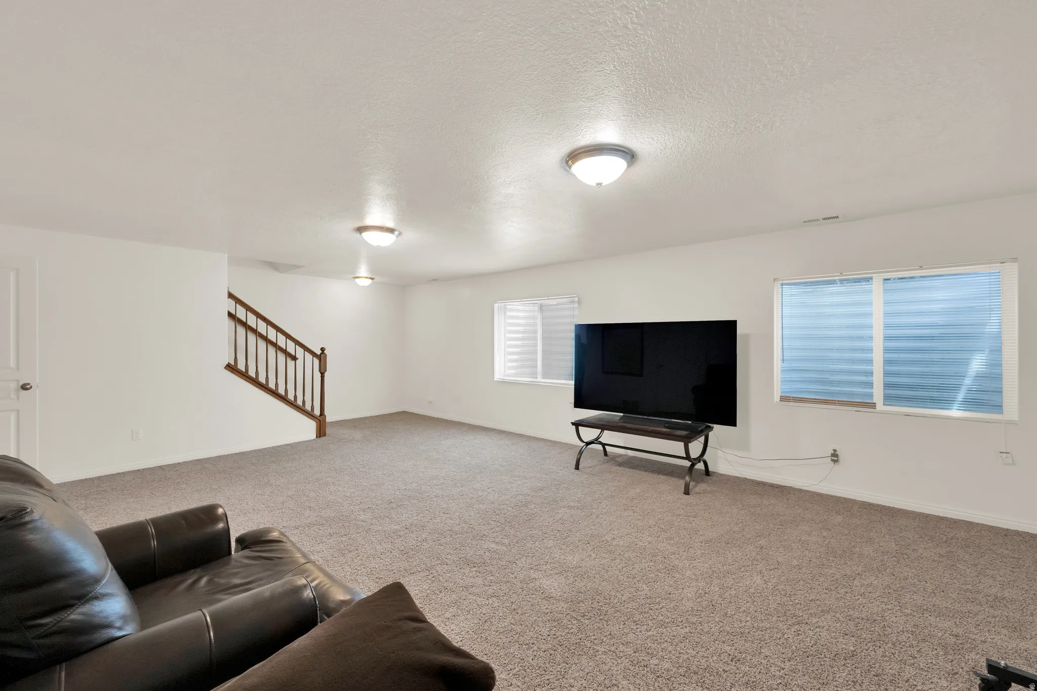 Spacious room featuring neutral carpet flooring, white walls, two windows with horizontal blinds, and a built-in wooden staircase with balusters